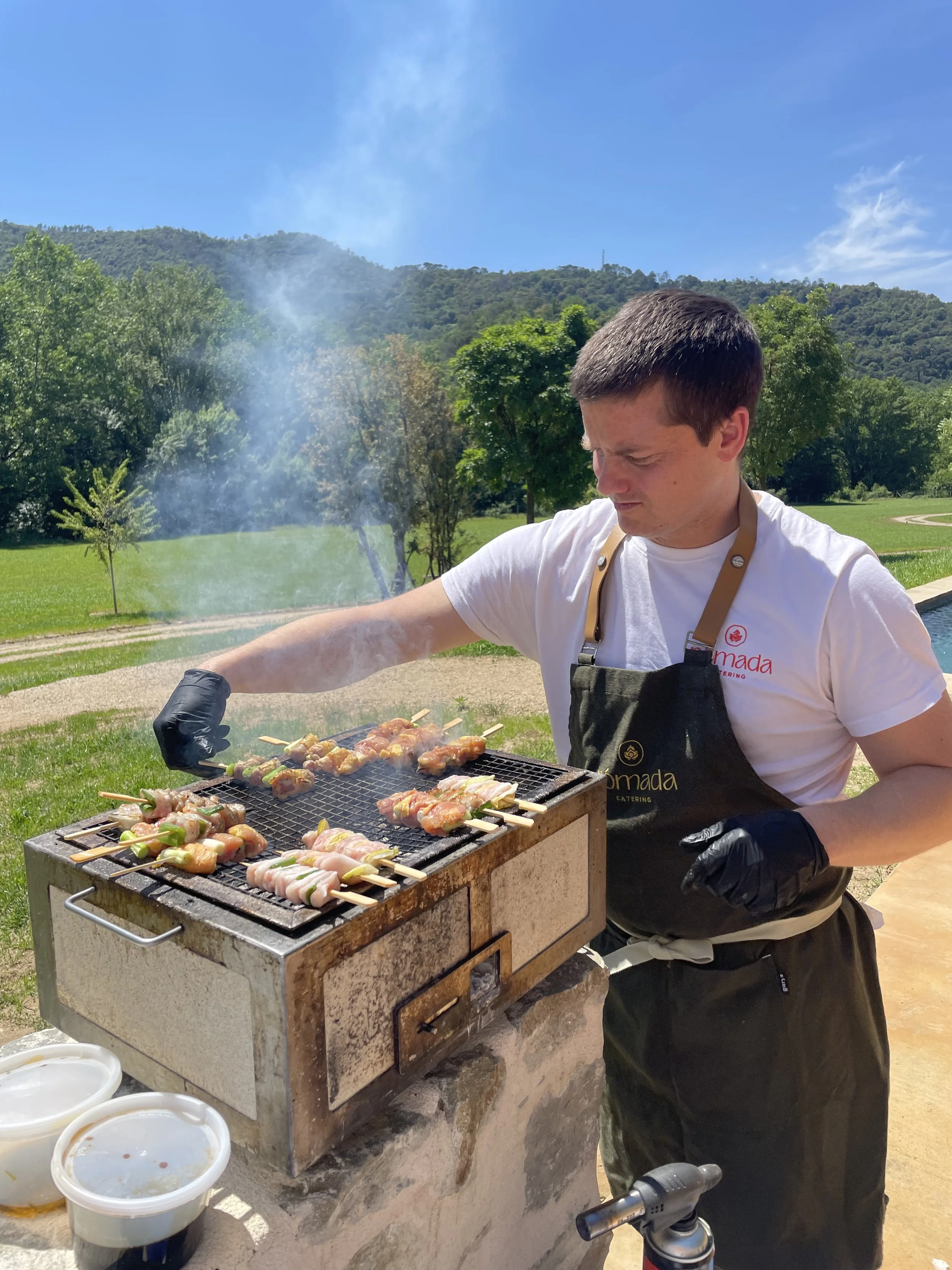 A man grilling skewers of food on an outdoor barbecue with a scenic background of green trees, grassy field, and hills under a clear blue sky.
