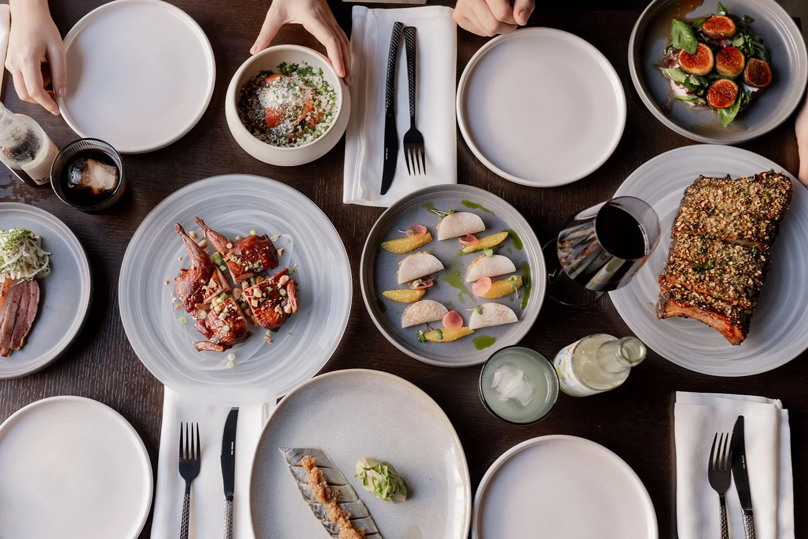 A table set with various dishes including a plate of glazed ribs, a small bowl of salad, an appetizer with white fish, a dish of sliced meat with garnish, a bowl of figs with greens, a glass of soda, and other plates with utensils and napkins.