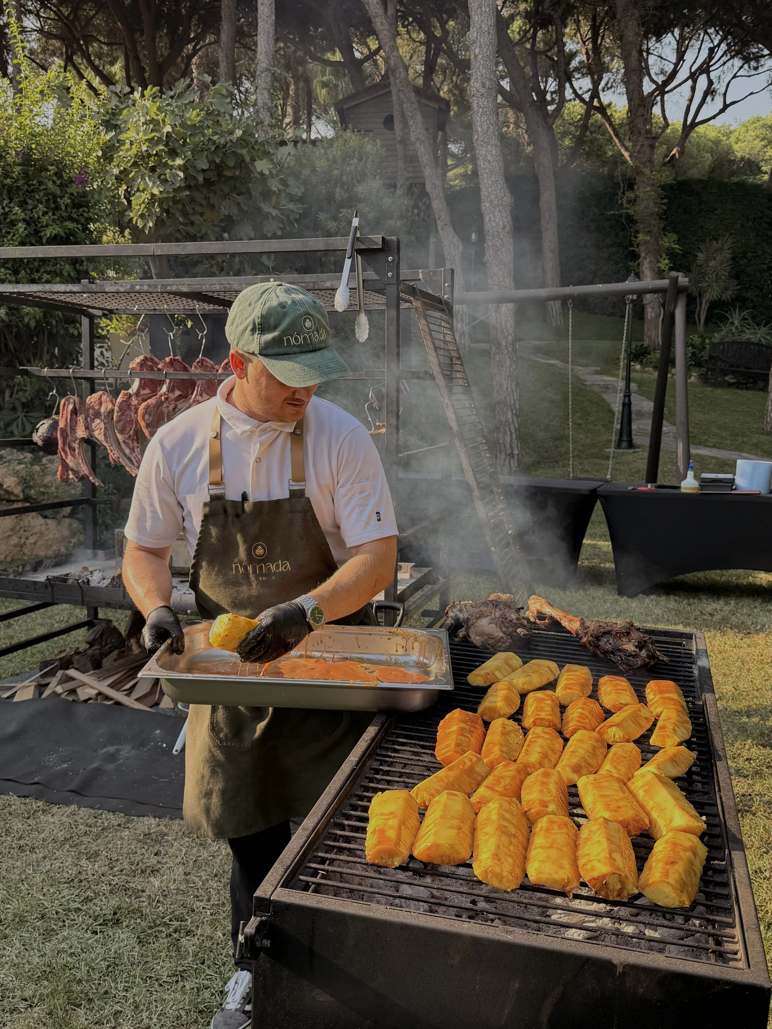 Man preparing food on an outdoor grill, with baked potatoes, grilled meats, and hanging cuts of meat in the background.
