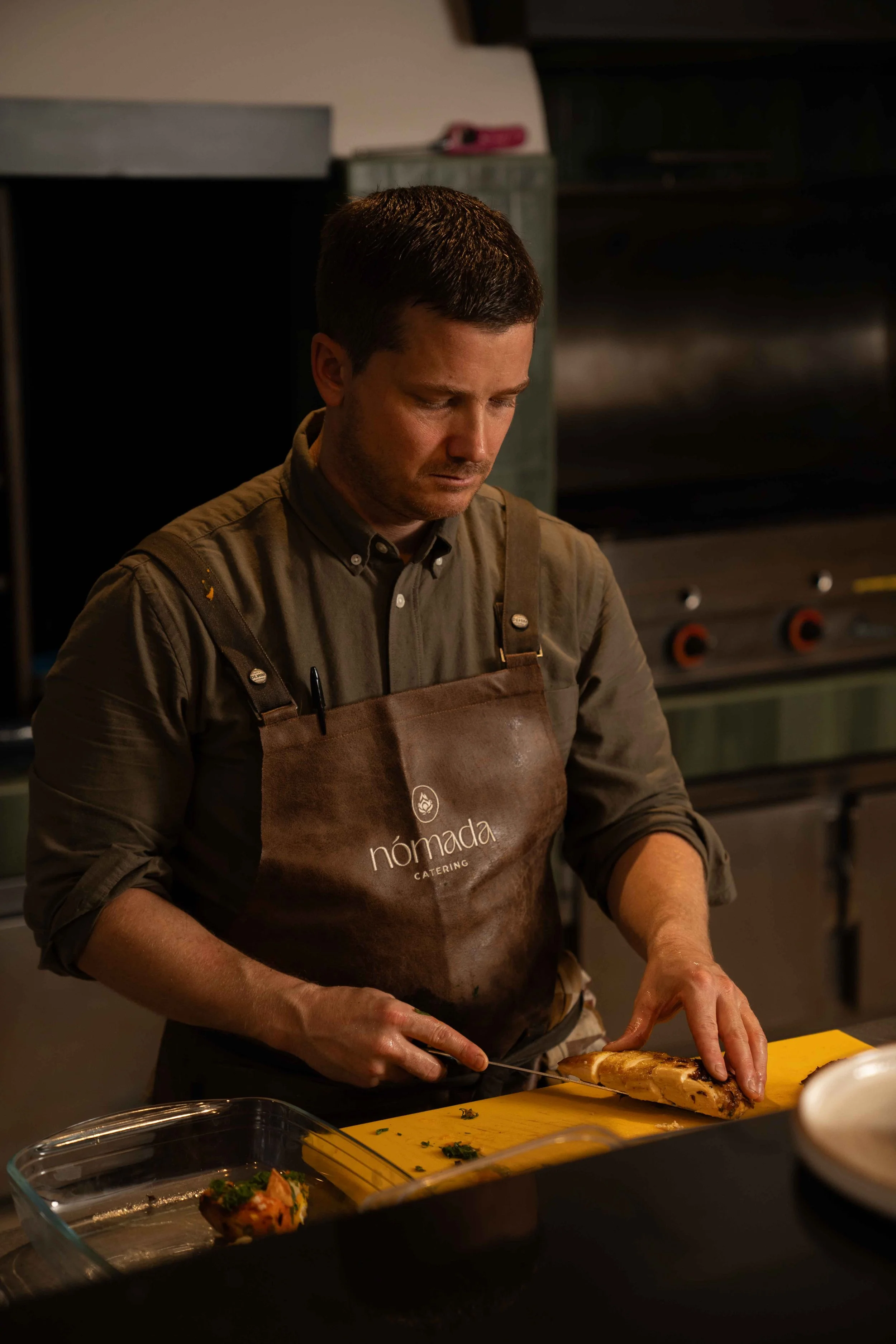 A man wearing a brown apron labeled 'Nomada Catering' is slicing a piece of roasted meat in a kitchen.