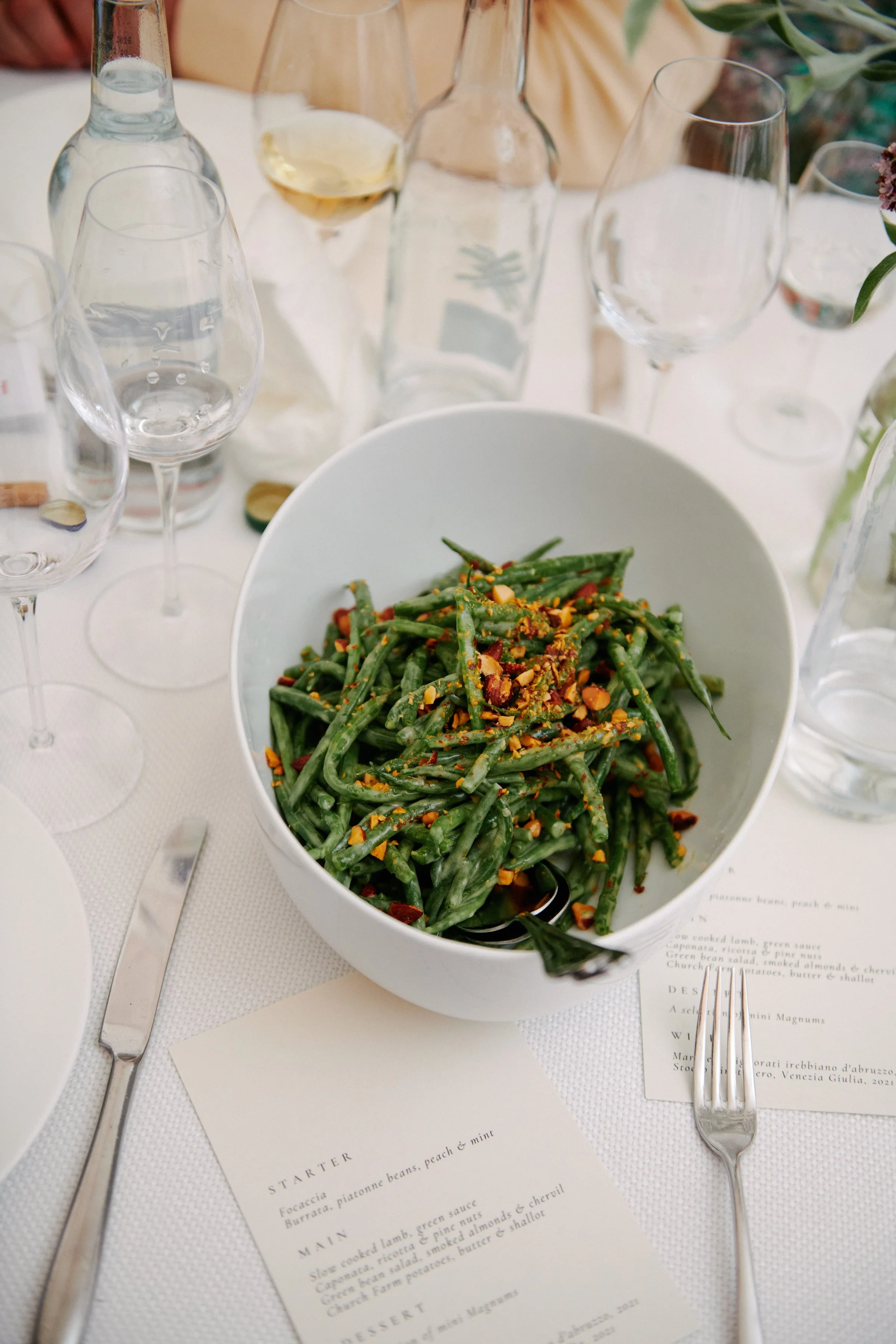 Green bean salad topped with chopped nuts served in a white bowl at a table with glassware and printed menu.