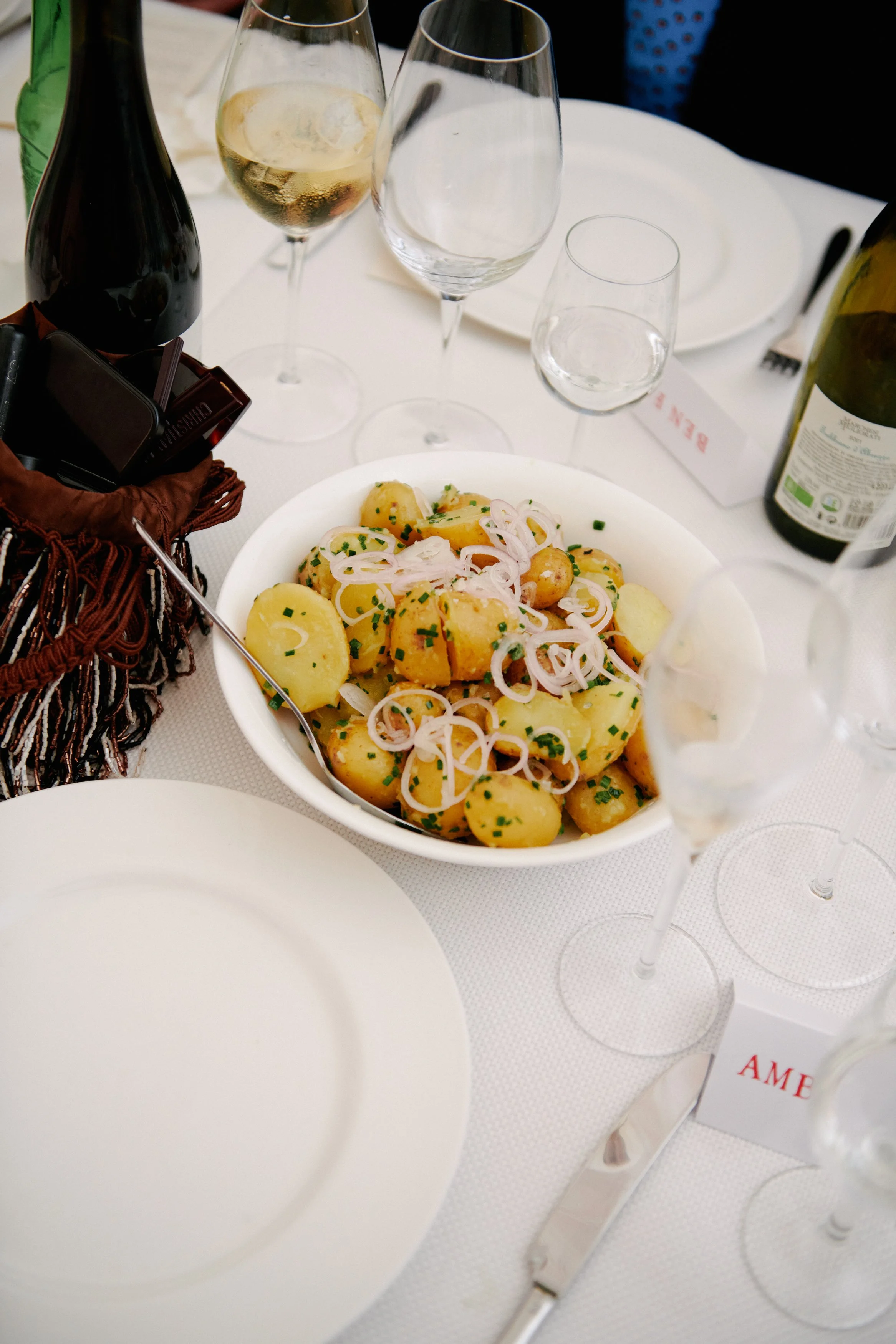 A table set for a meal with a large bowl of cooked potatoes garnished with chopped chives and sliced onions, surrounded by empty and partially filled wine glasses, bottles of wine, and white plates on a white tablecloth.