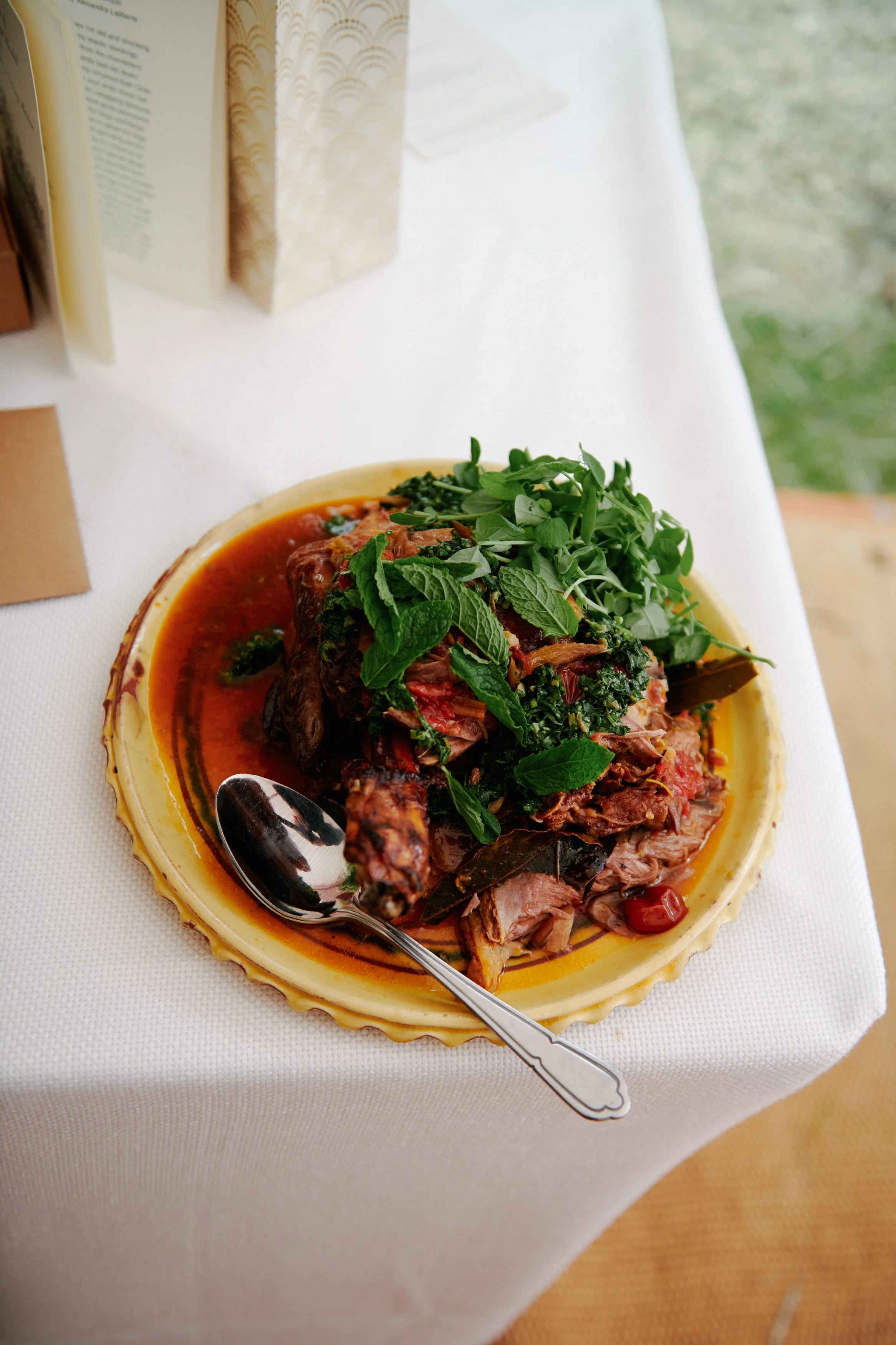 A plated cooked meat dish garnished with fresh herbs and vegetables, served on a white tablecloth.