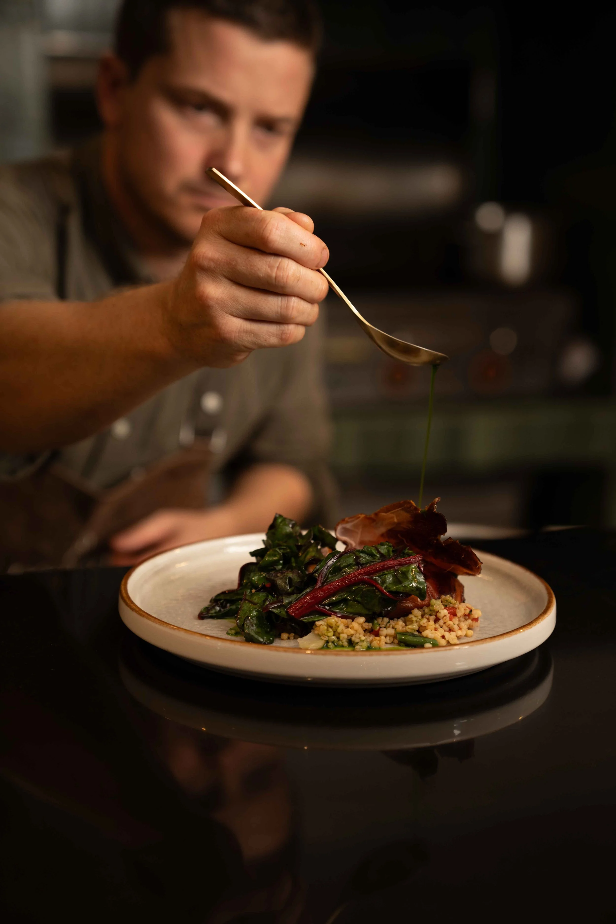 A chef drizzling olive oil over a plated gourmet meal that includes greens and grains in a professional kitchen.