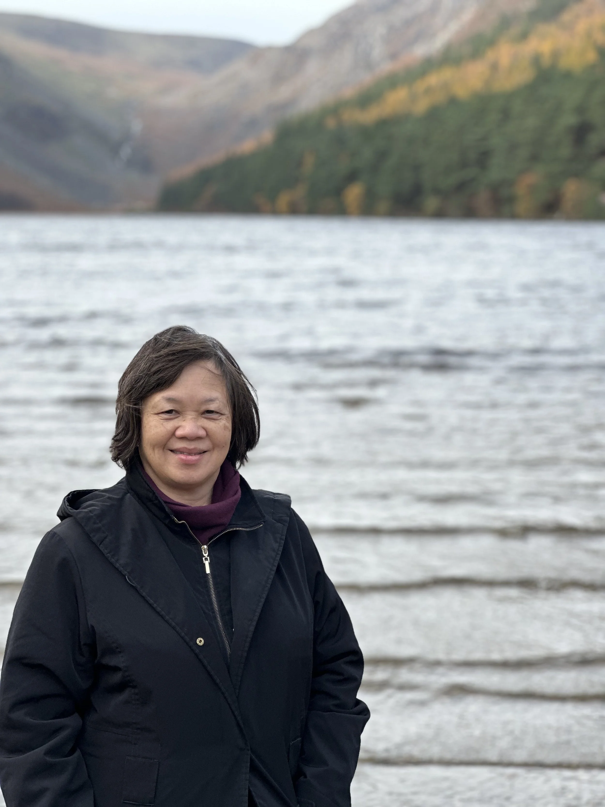 A woman with short dark hair smiling, wearing a black jacket, standing by a lake with mountains in the background.