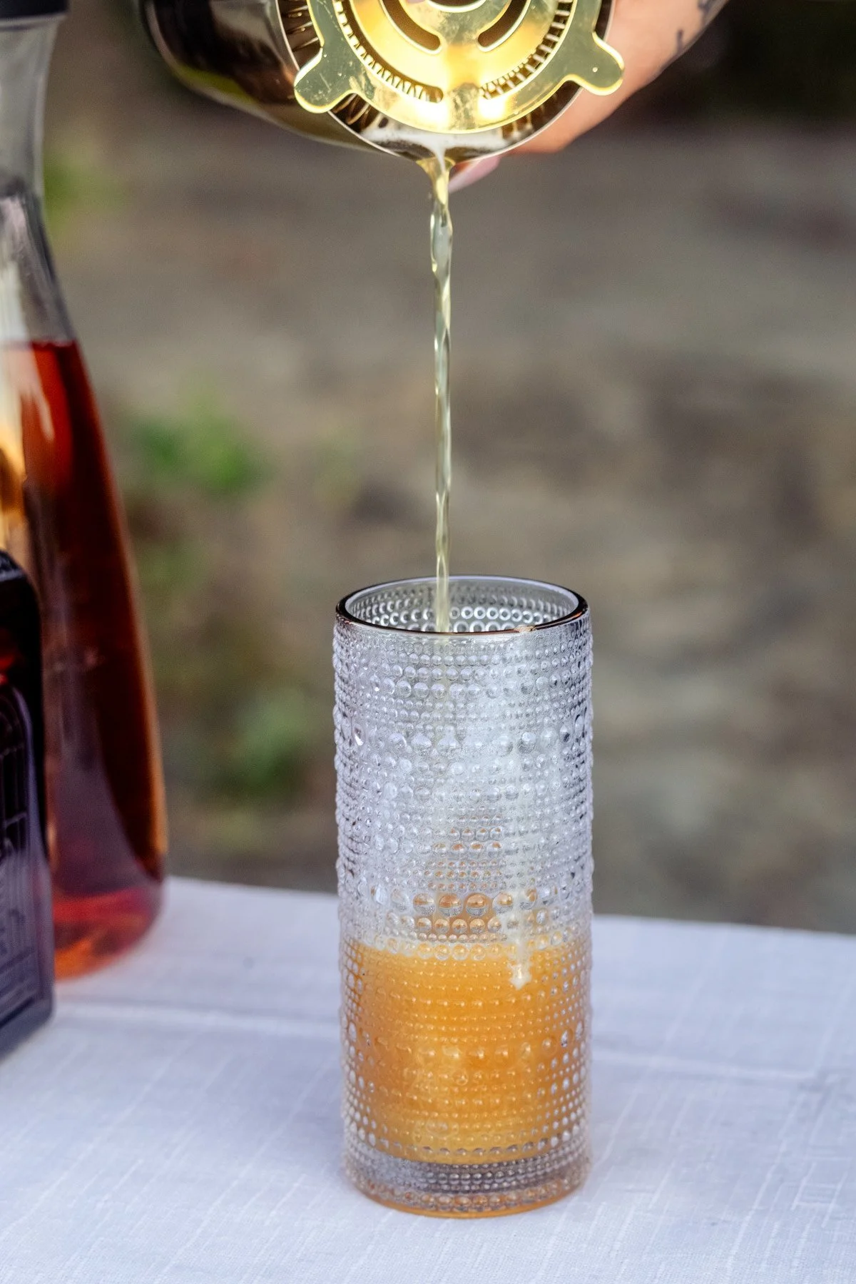 A hand pouring beer from a green bottle into a textured glass on a table.