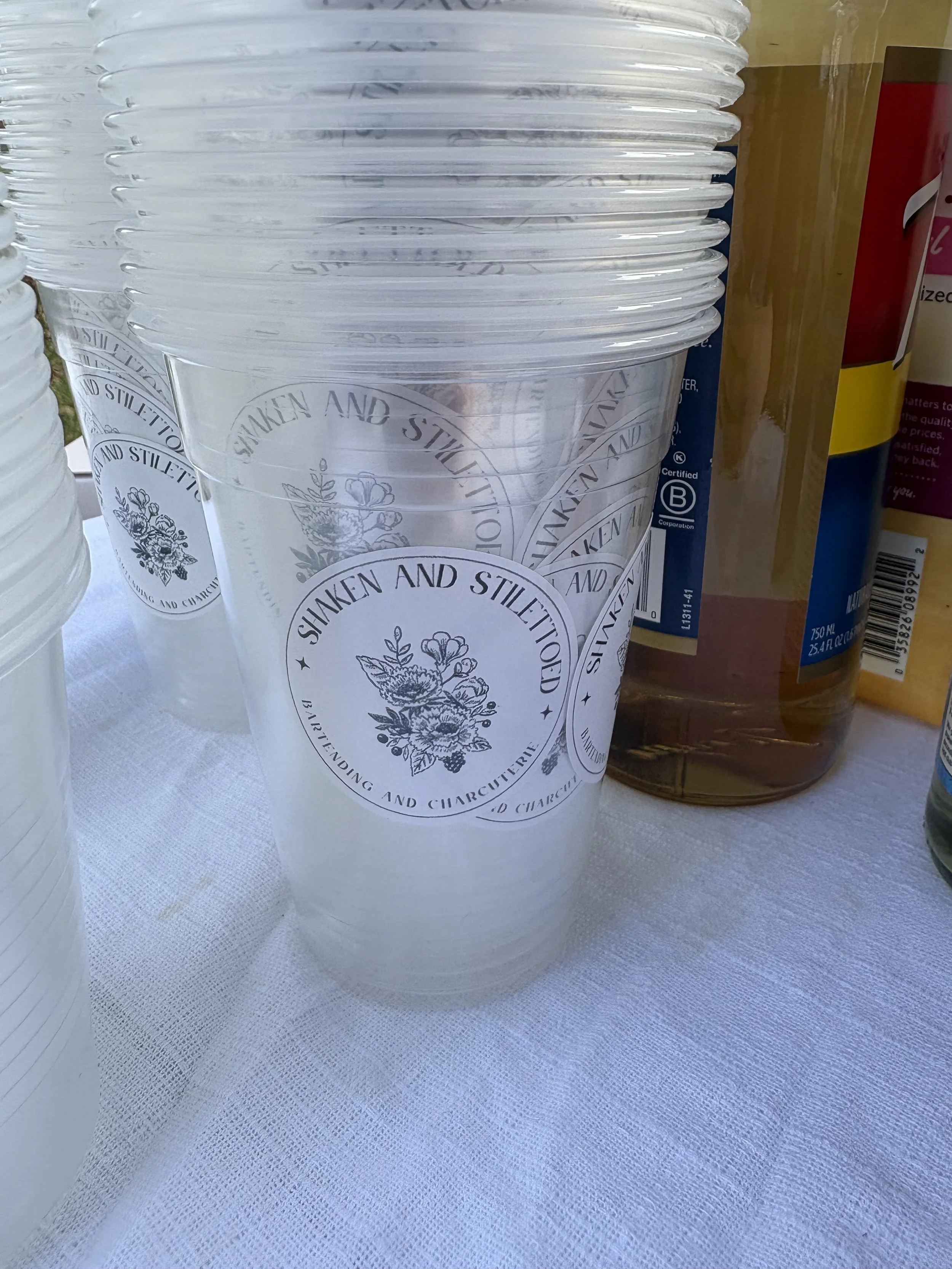 Clear plastic cups with a black and white floral logo and the text "Shaken and Stil COVID," stacked on a white tablecloth.