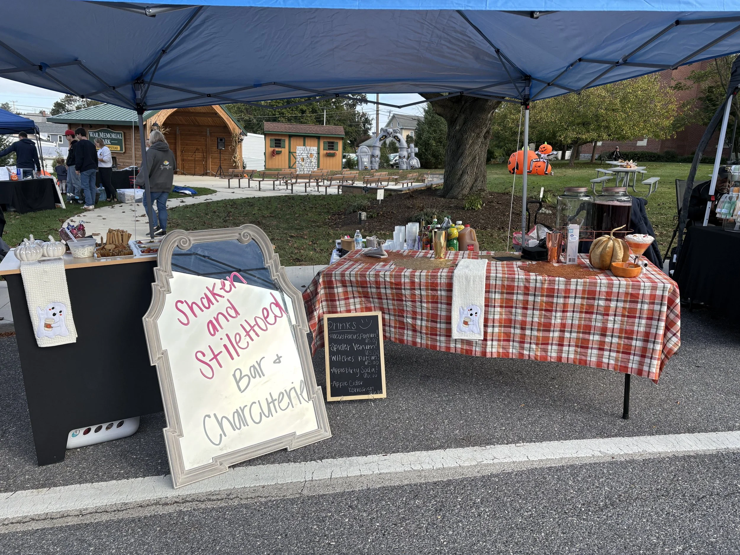 Market stall with a sign reading 'Shaken and Stilted Bar & Charcuterie,' under a blue canopy. The stall has a plaid tablecloth, pumpkins, drinks, and snacks. Autumn decorations and Halloween-themed items are visible, with a park and people in the background.