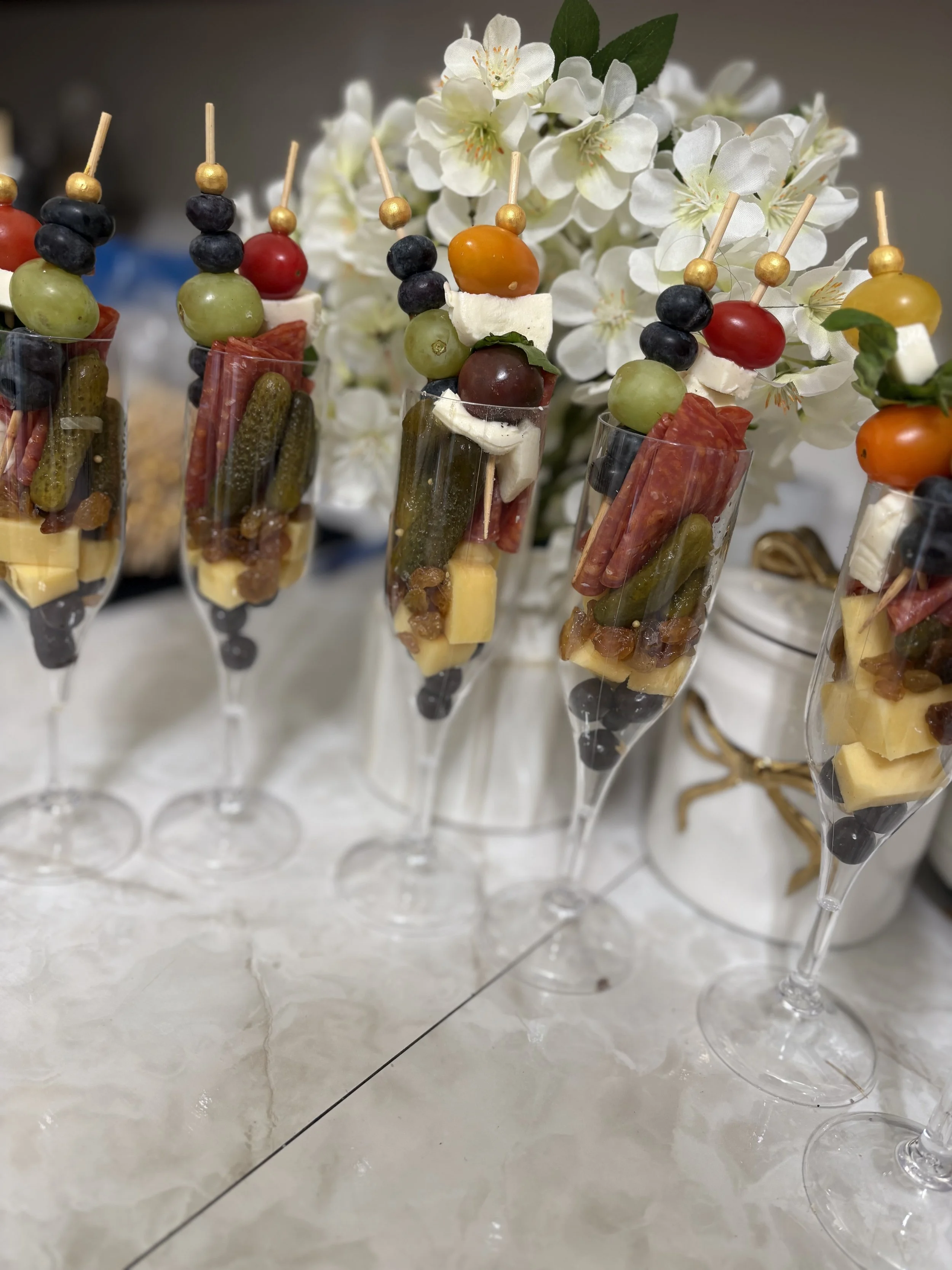 Champagne glasses filled with layered cheese, meats, pickles, and grapes, garnished with skewers topped with various cheeses, grapes, cherry tomatoes, and cherry peppers, with white flowers in the background.