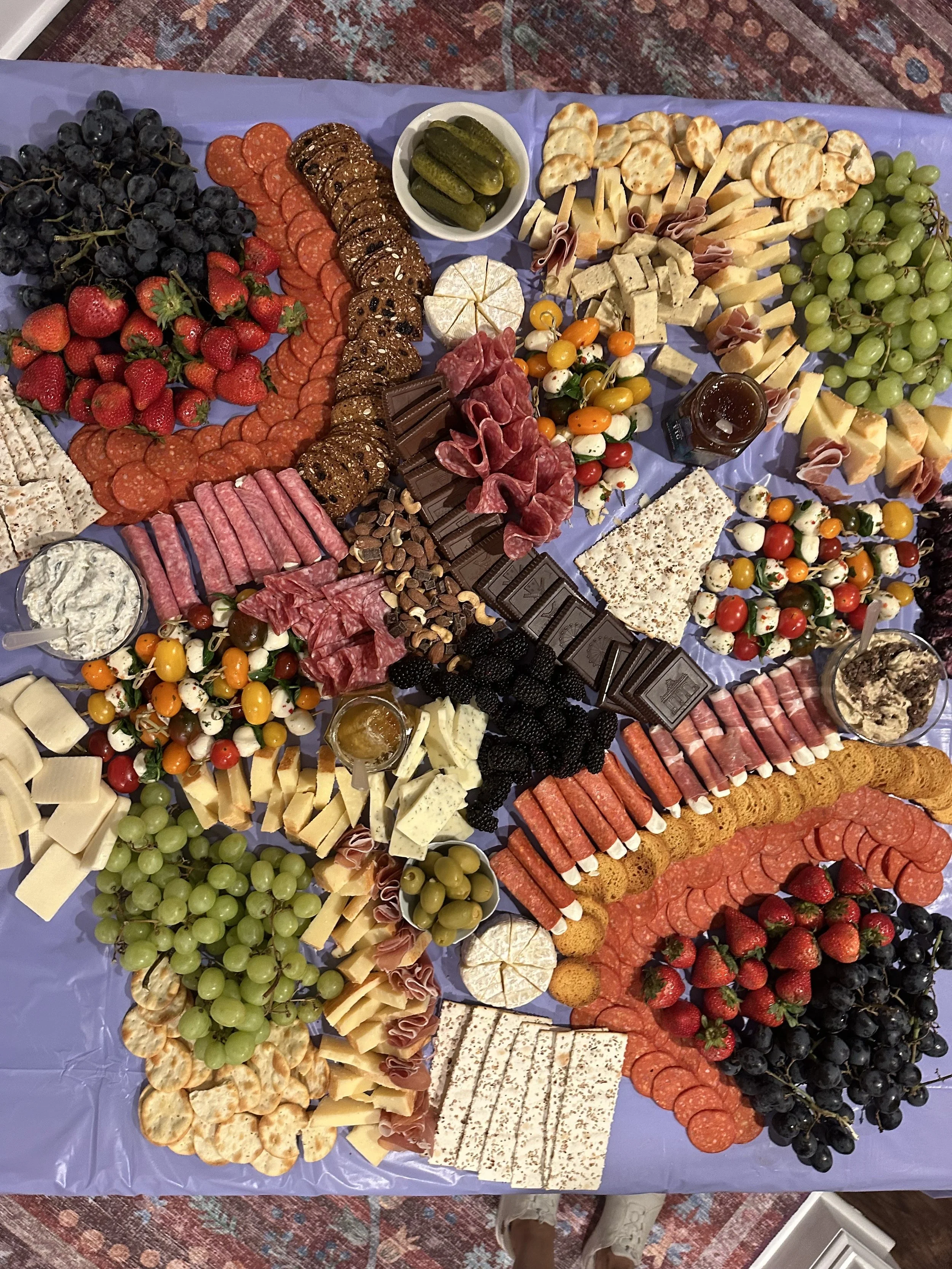 A large charcuterie and fruit platter arranged with strawberries, grapes, blackberries, cherry tomatoes, various cheeses, sliced meats, crackers, pickles, chocolate, jams, and spreads on a purple tablecloth.
