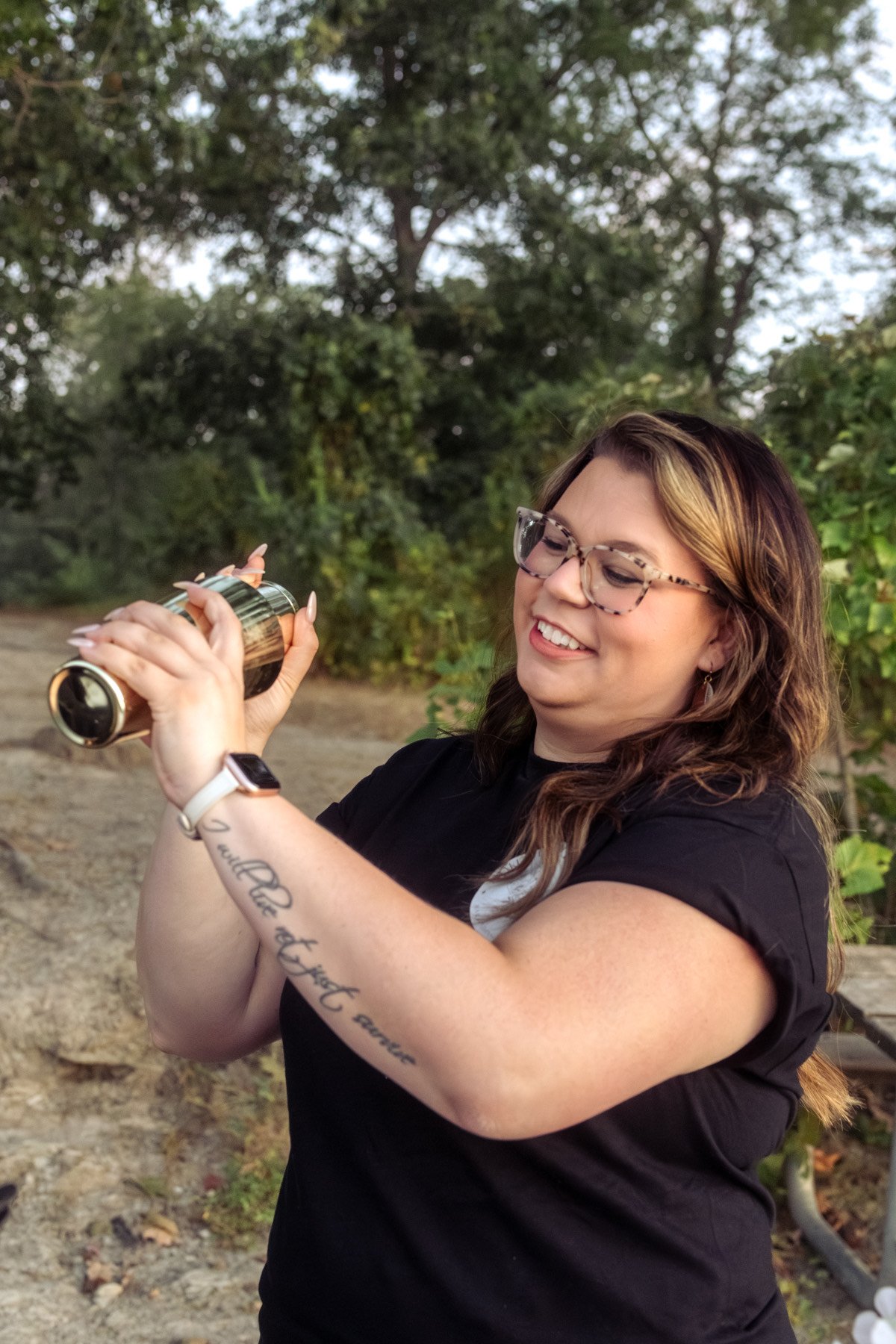A woman with glasses and tattoos is smiling and holding a metallic thermos outdoors, with trees and greenery in the background.