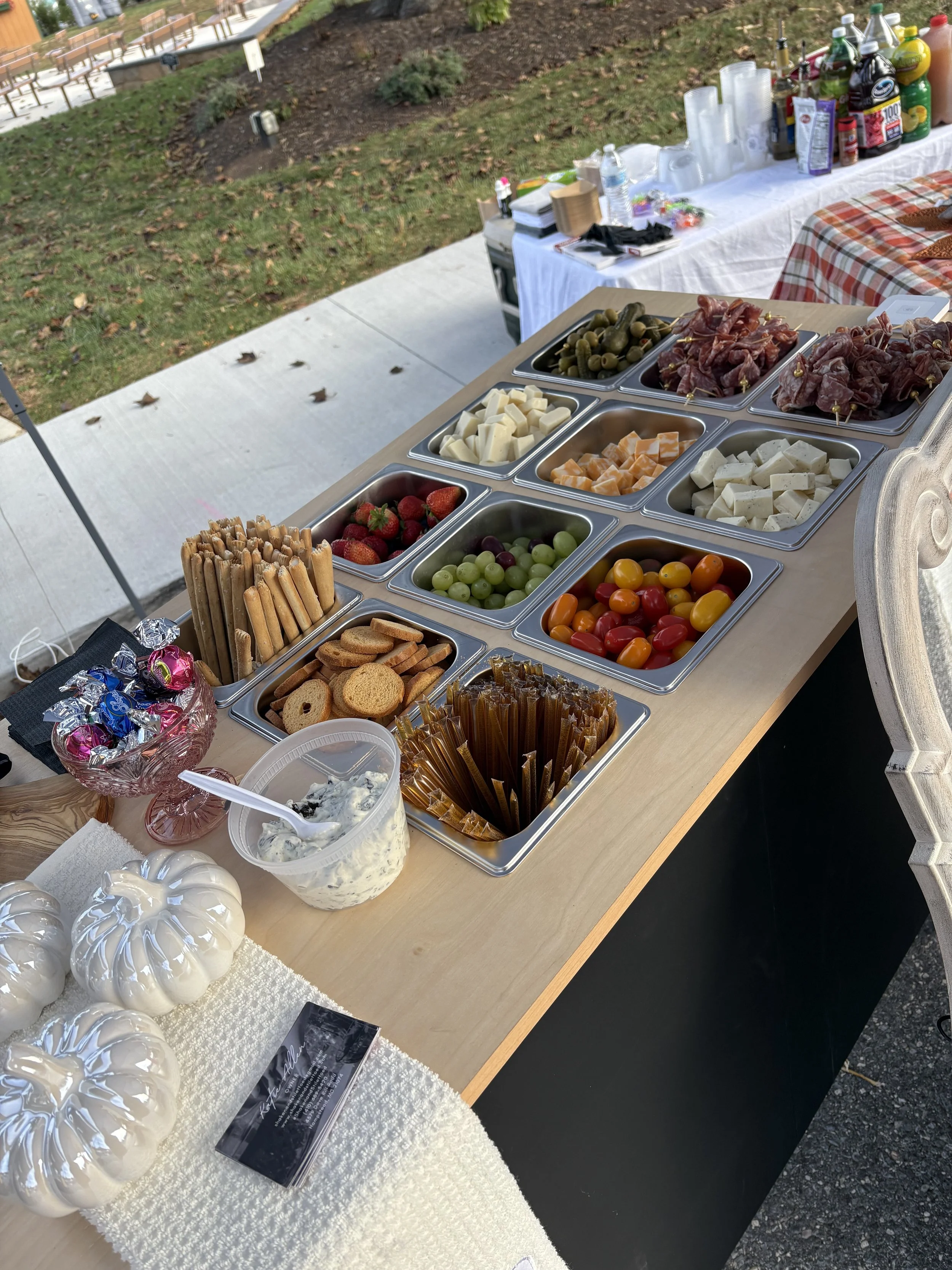A buffet table outdoors with a variety of foods including cheese, strawberries, green and purple grapes, cherry tomatoes, crackers, breadsticks, wrapped candies, dips, and cooked meats, with soda bottles and cups in the background.