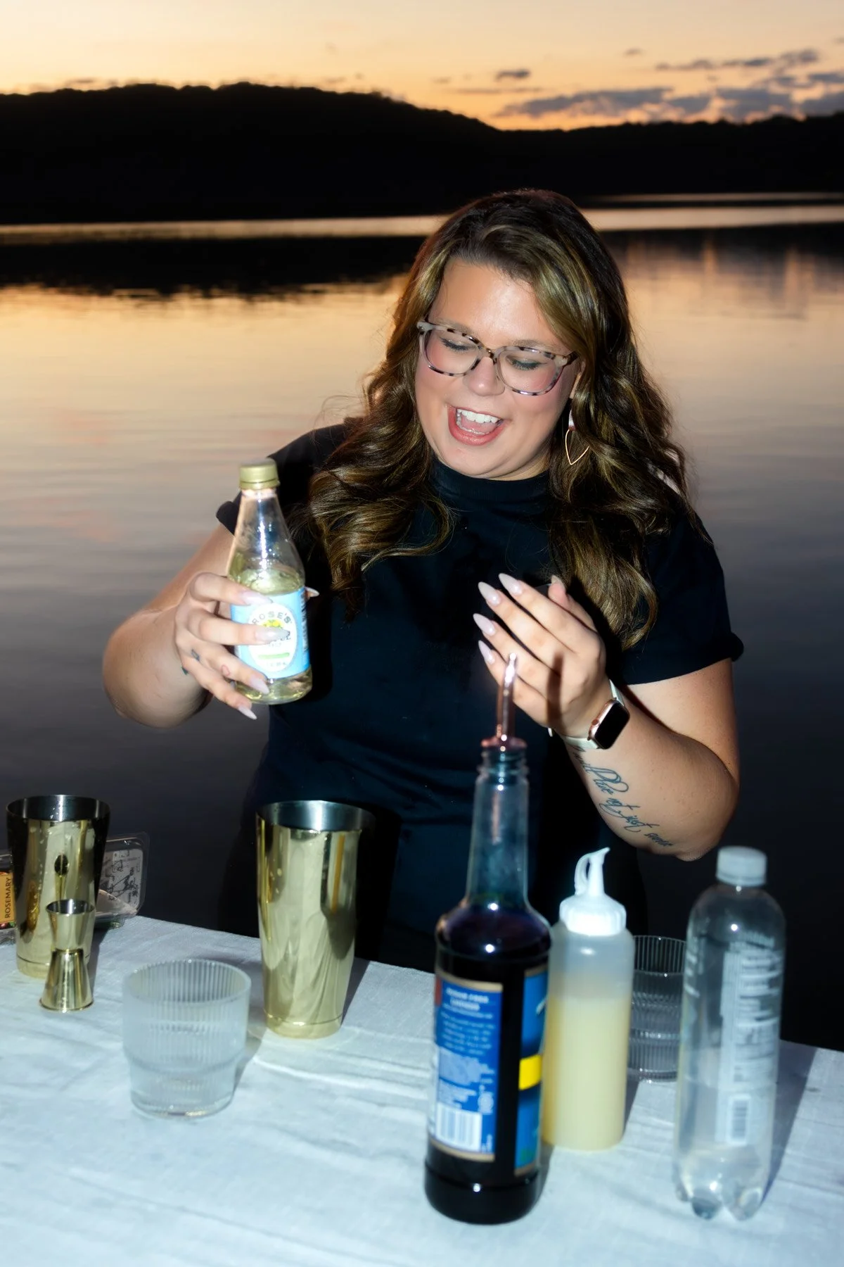 A woman with long wavy hair and glasses holding a bottle of sparkling water, celebrating outdoors at sunset with drinks and a table set up by a lake.