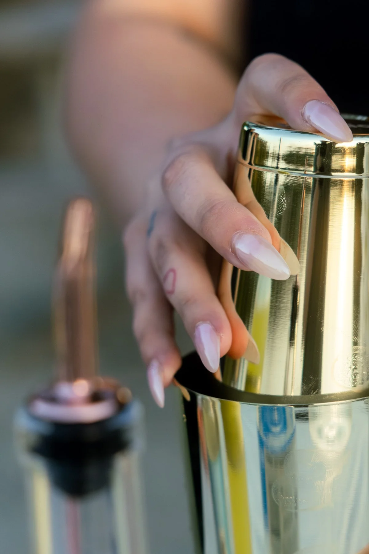 Close-up of a woman's hand with long manicured nails holding a shiny gold trophy cup, with a blurred perfume bottle in the foreground and a colorful outdoor background.