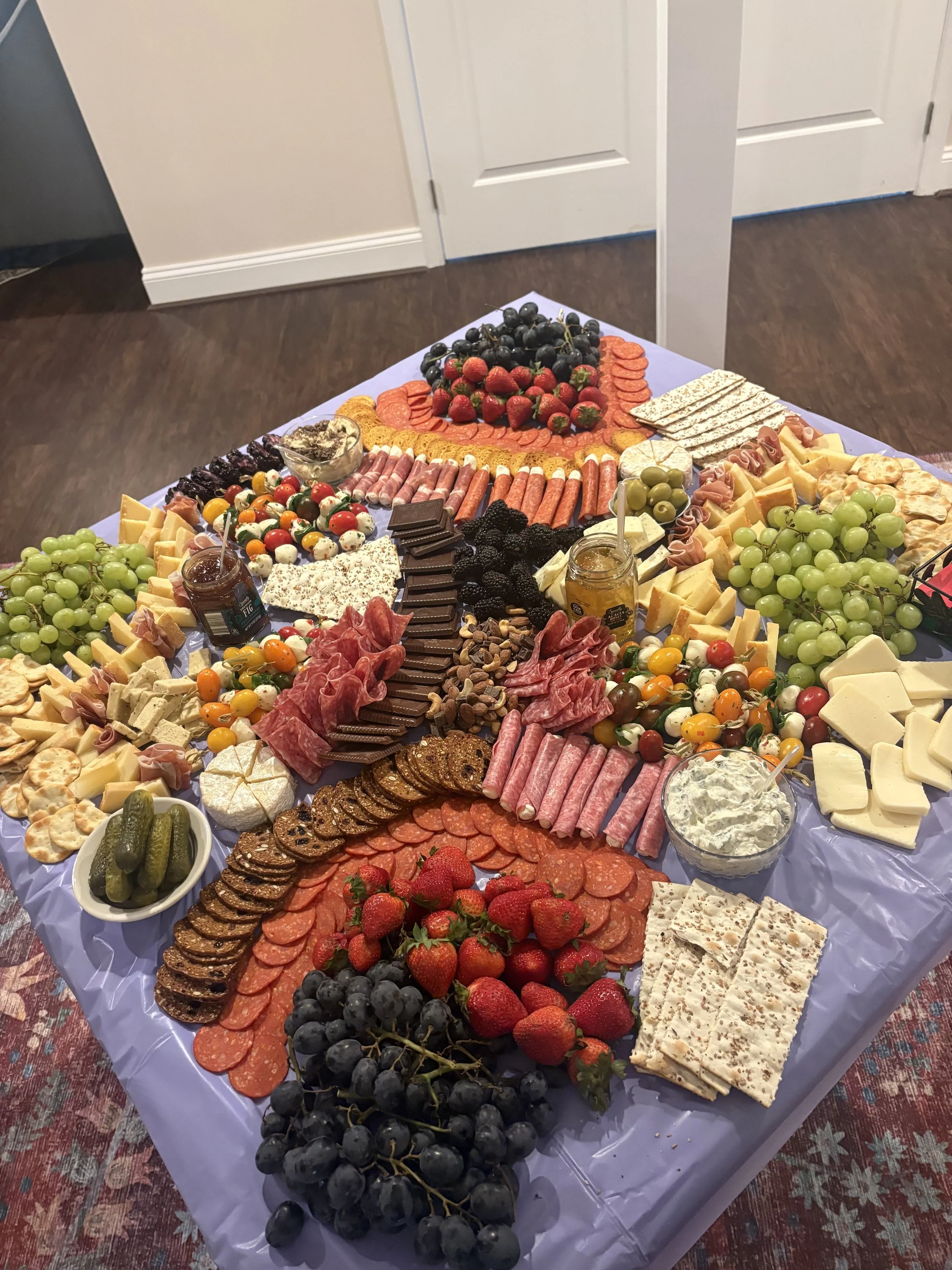 A large table covered with a variety of cheeses, sliced meats, fruits, crackers, and condiments arranged for a buffet or party setting.
