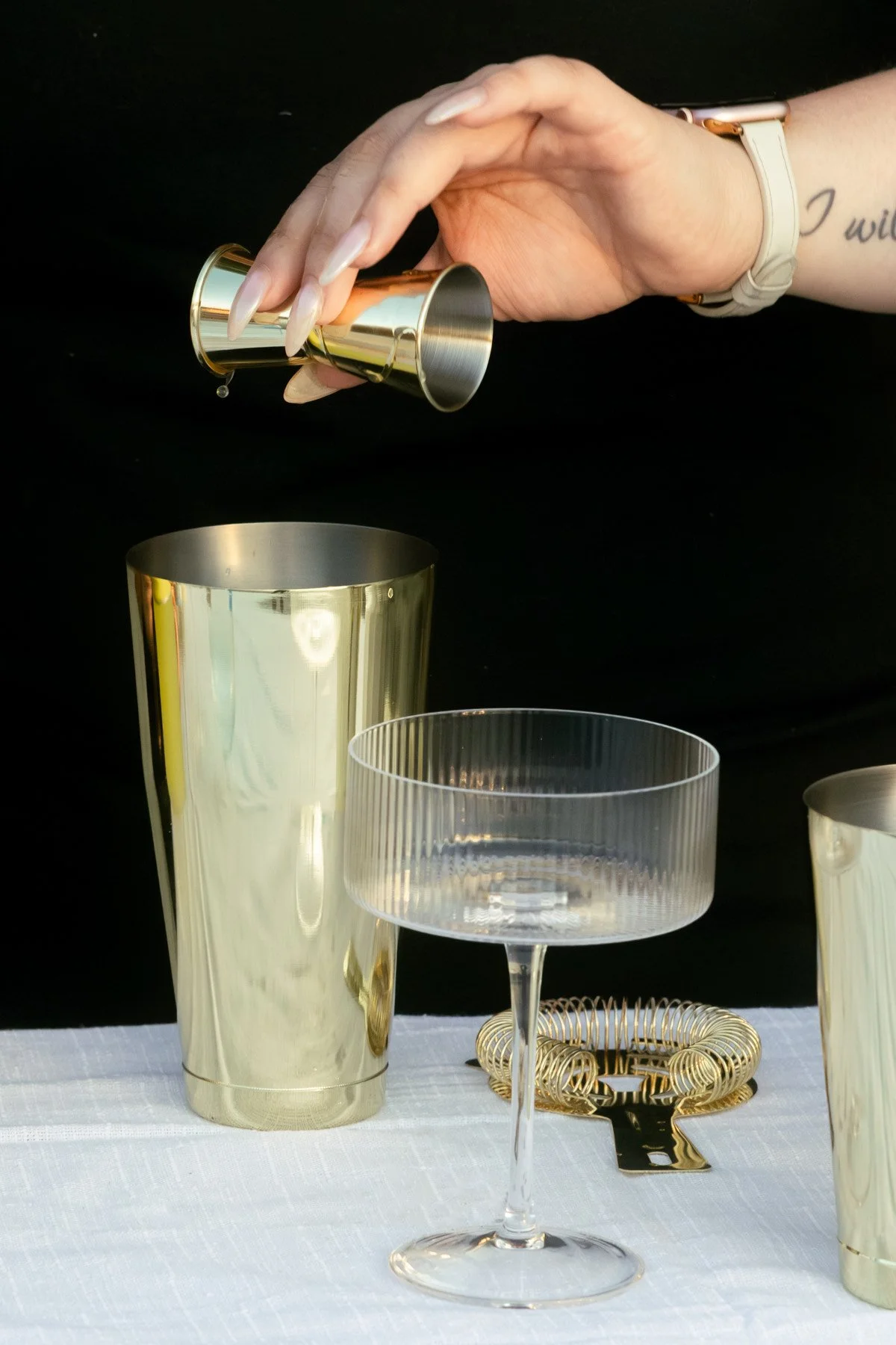 Person pouring a drink into a cocktail glass with gold and silver barware on a white tablecloth against a black background.