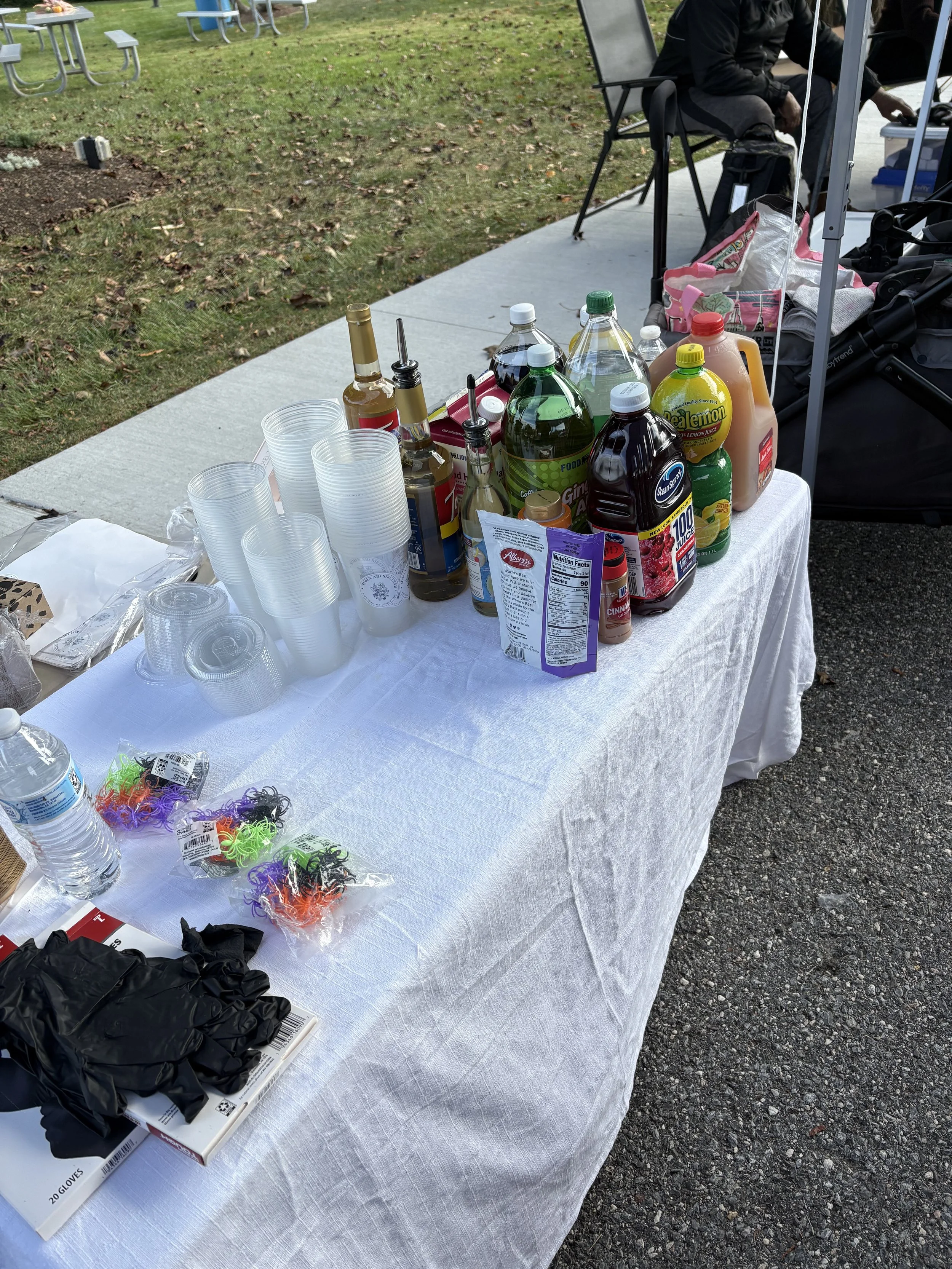 A table outdoors with various bottles of drinks, plastic cups, and small colorful hair ties. There are some black gloves and a book on the table, and a person sitting in the background.