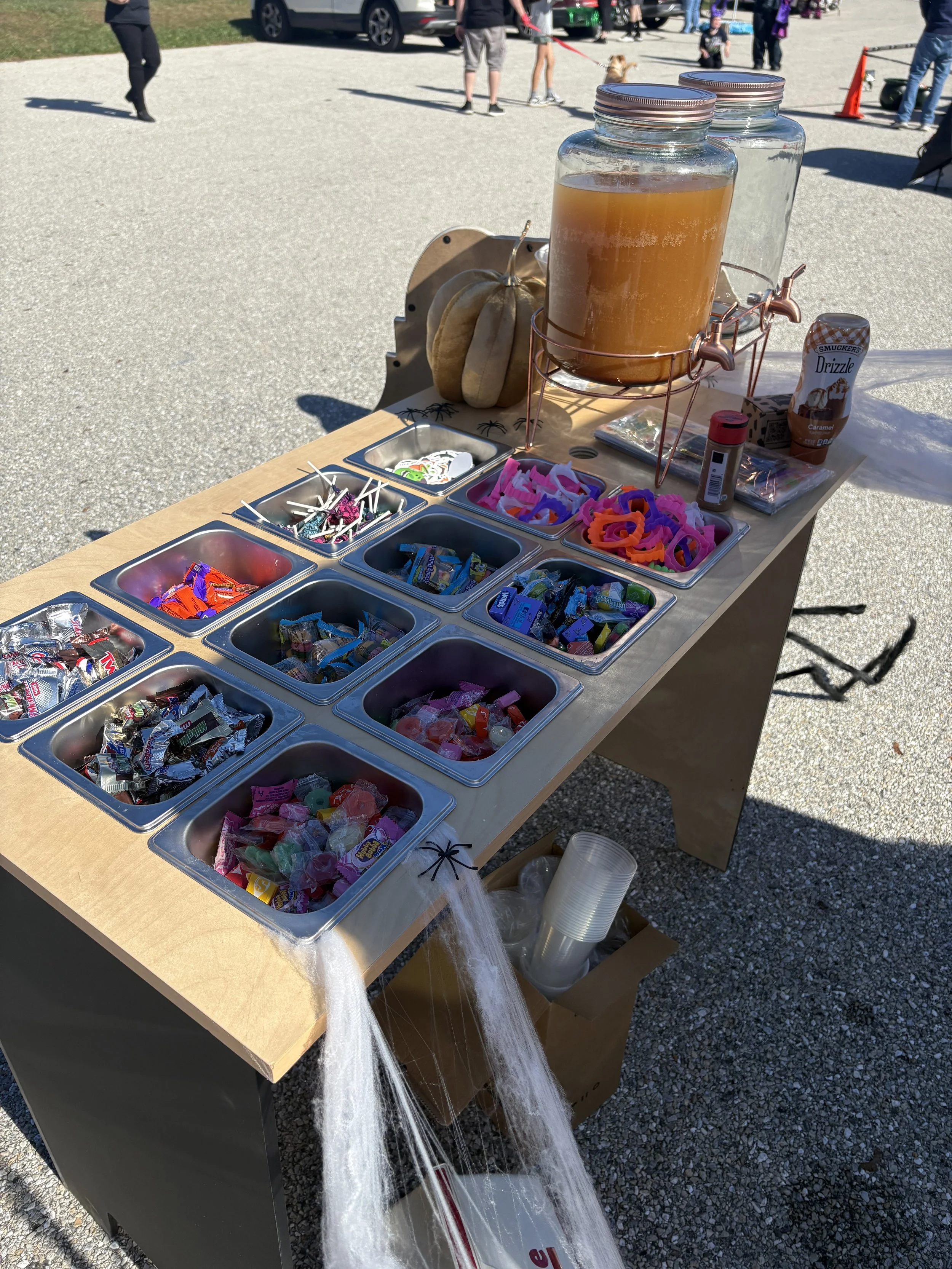 Candy station at an outdoor event with jars of drinks and various candies in metal containers, surrounded by people.