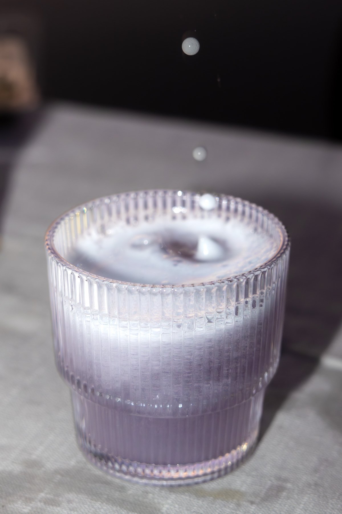 A clear, textured glass tumbler filled with water and ice cubes, placed on a light-colored surface. Droplets are falling into the drink, creating ripples.