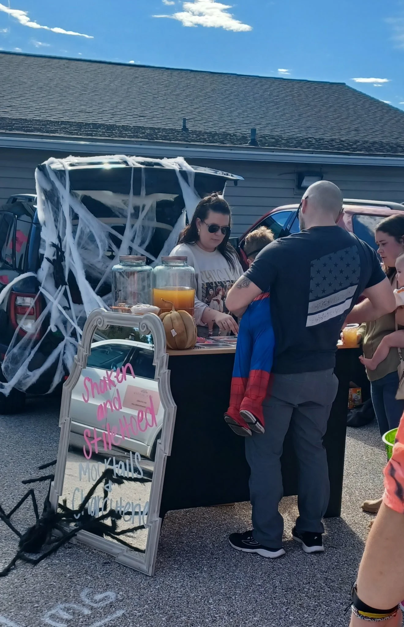 People gathered around a Halloween-themed snack stand decorated with fake cobwebs, pumpkin, and jars of juice at an outdoor event.