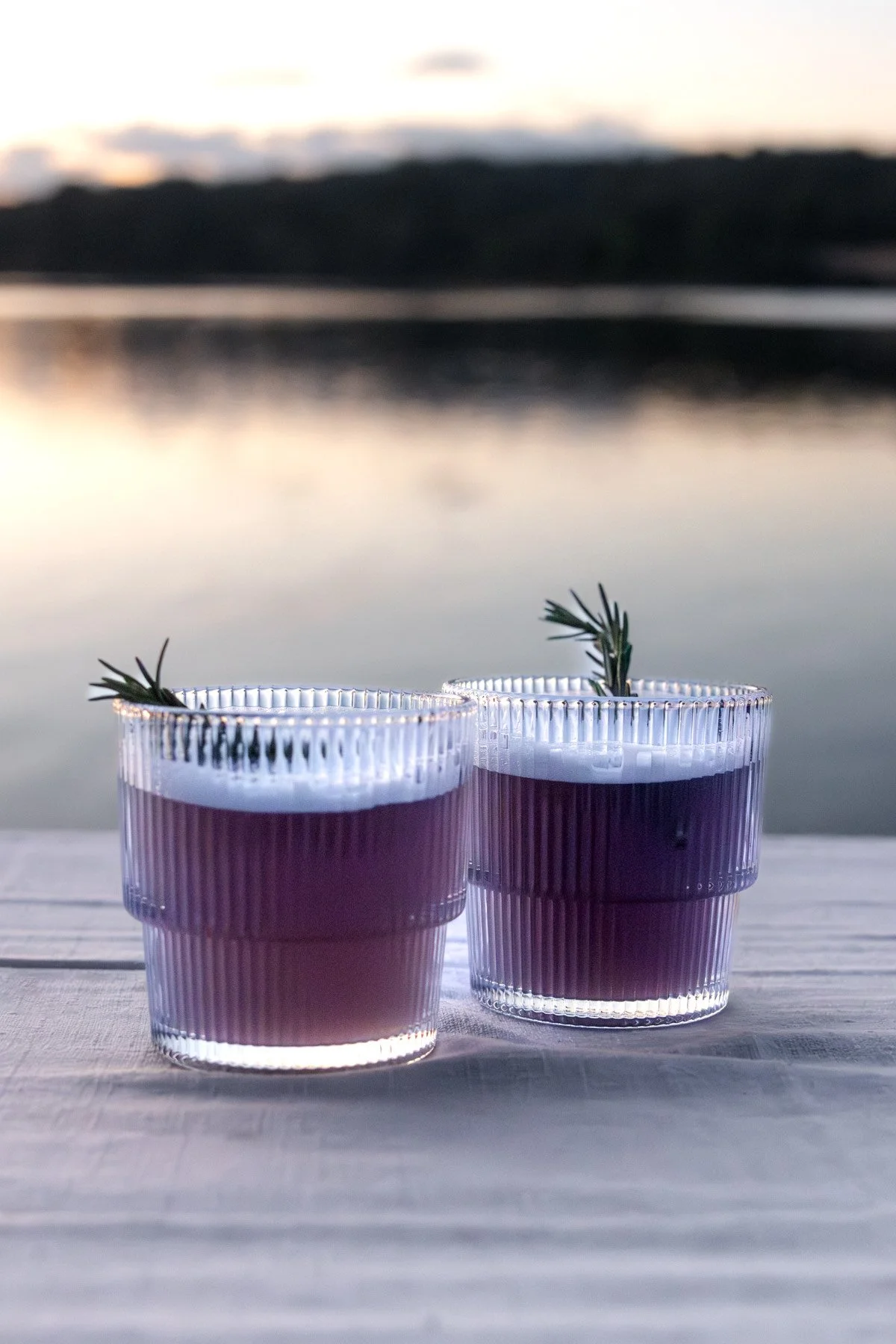Two glasses of dark purple beverage garnished with sprigs of rosemary on a wooden surface outdoors at sunset, with a blurred lake and mountain silhouette in the background.