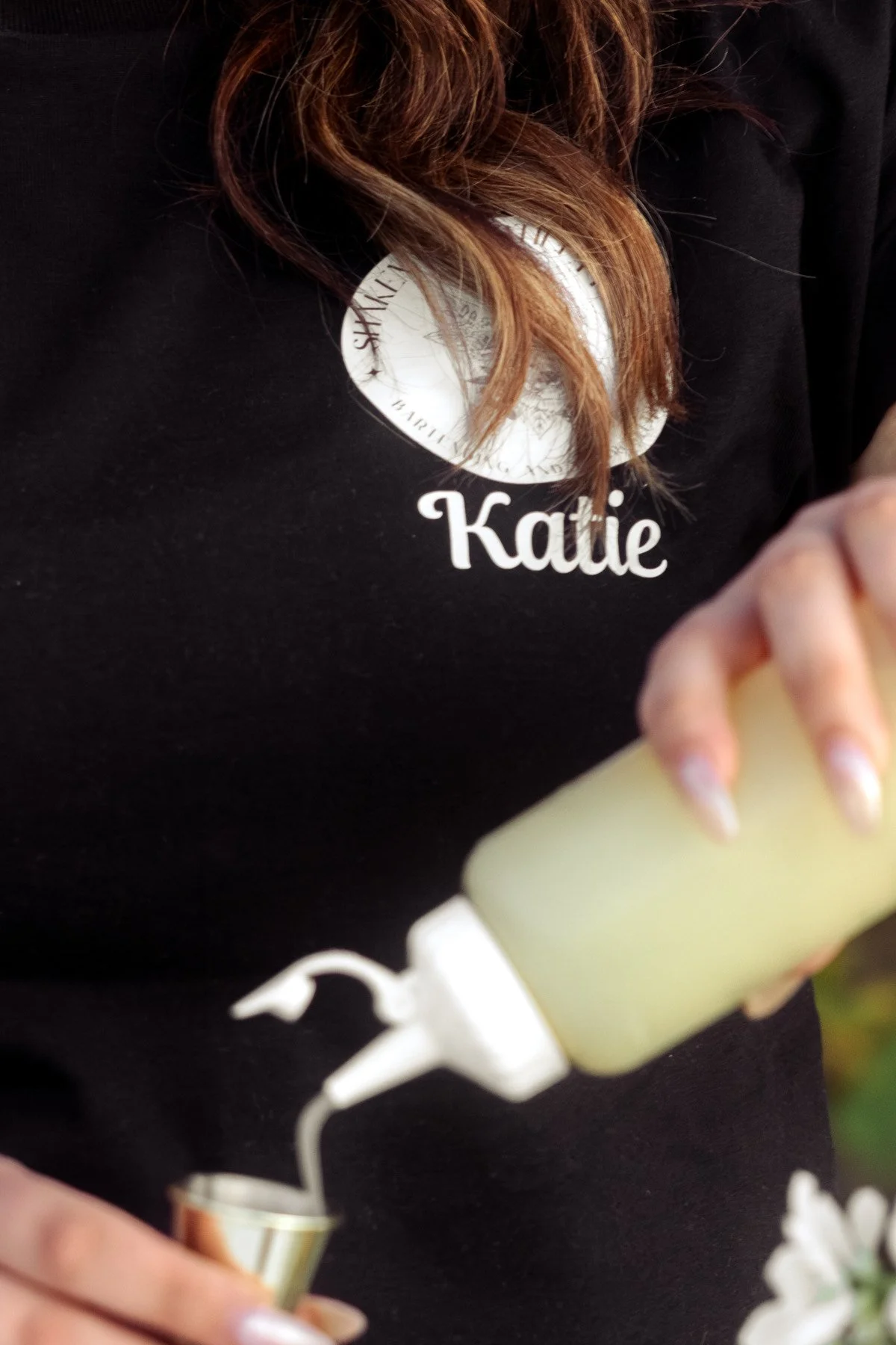 A woman with brown hair in a black shirt with a name badge that reads "Katie" is pouring a yellowish beverage from a squeeze bottle into a small cup.