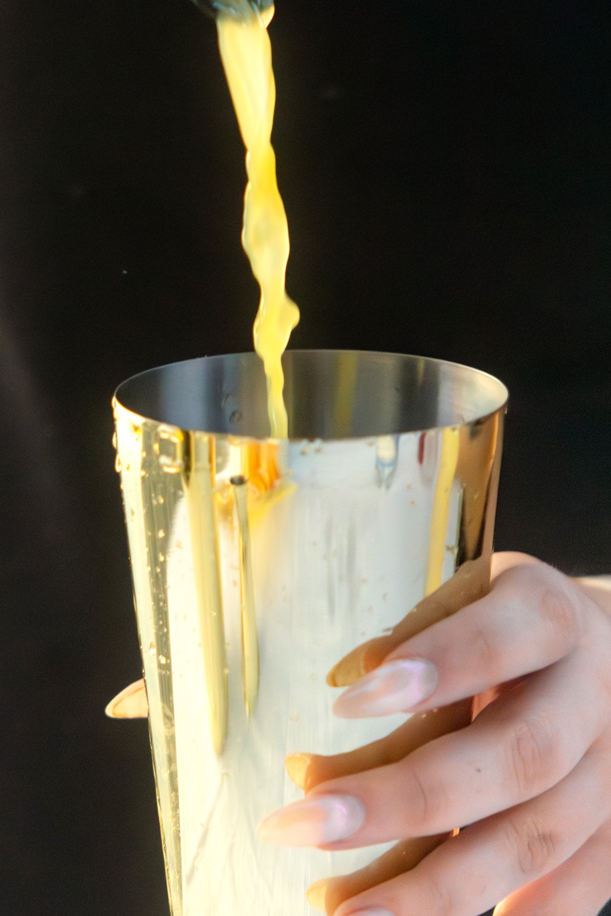 A hand holding a shiny metal cup with yellow liquid being poured into it against a dark background.