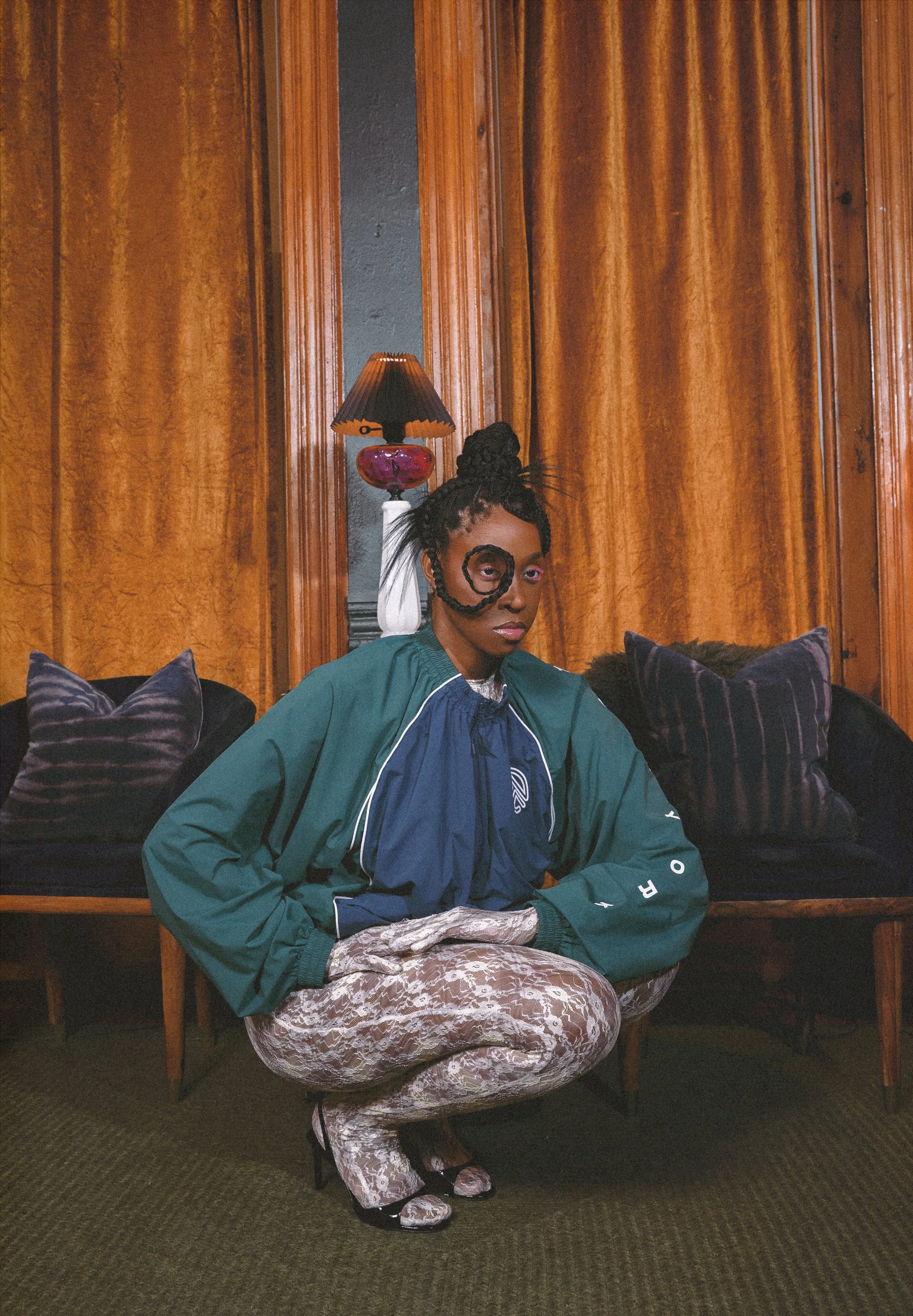A woman with an elaborate hairstyle, wearing a multicolored jacket and patterned gold pants, is sitting cross-legged on a chair. She is in a room with wood-paneled walls, black velvet cushions, and a lamp with a dark shade and purple base in the background.