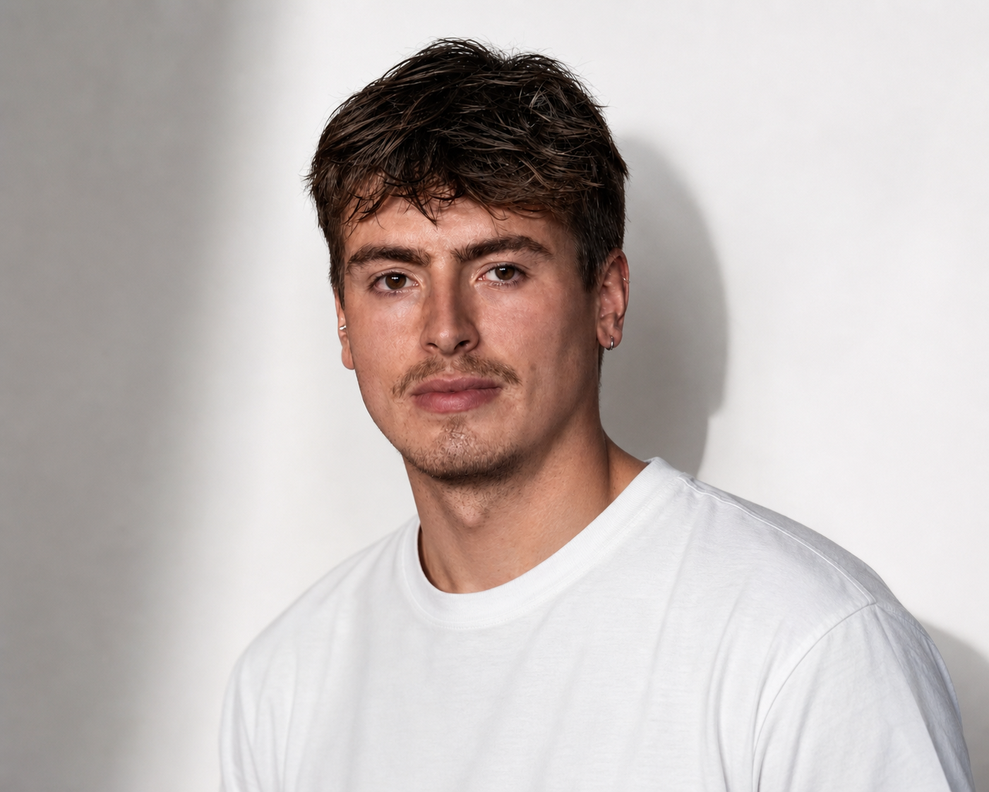 A young man with brown hair, a light mustache and goatee, wearing a white t-shirt, looking at the camera against a plain, light-colored background.