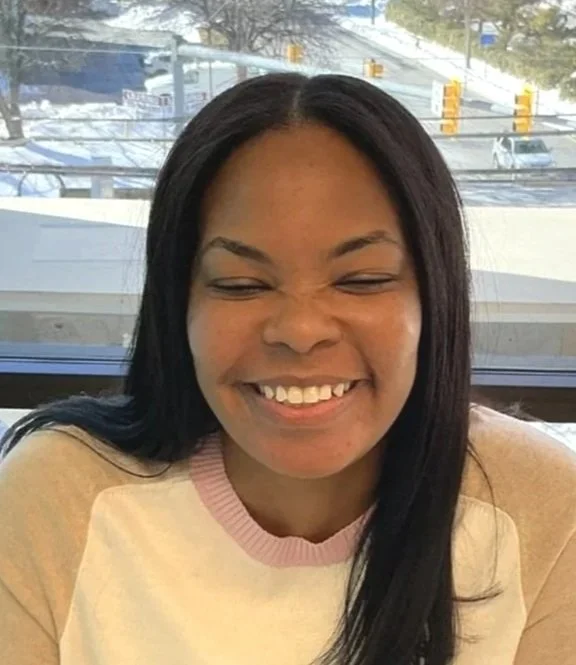 A woman with curly dark hair sitting indoors with a turquoise wall in the background, featuring a framed black and white photo of a young boy, and a side lamp illuminating her face.