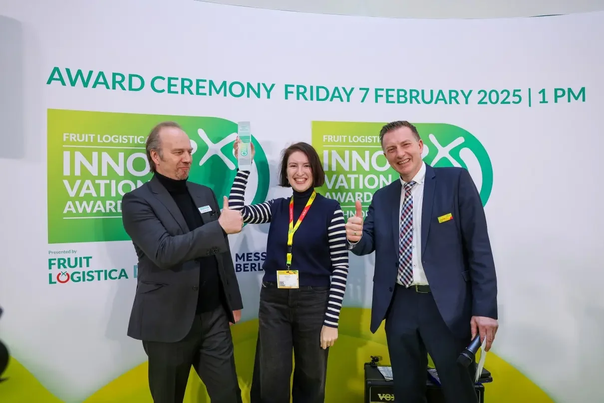 Three people posing with thumbs up at the Fruit Logistics Innovation Award ceremony held on February 7, 2025, in Berlin, with a backdrop displaying event details.