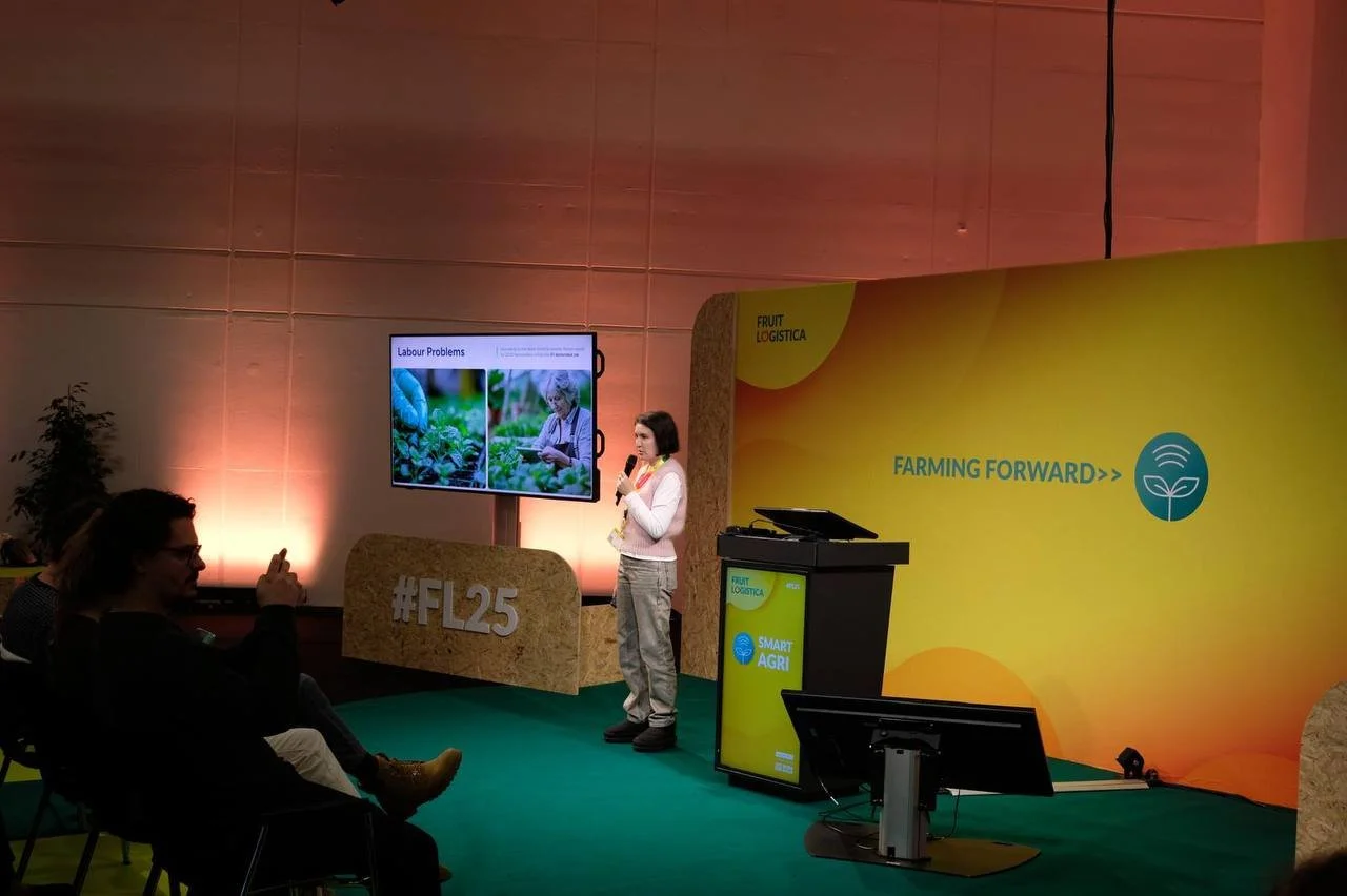 Woman giving a presentation on labor problems at a conference, with a large screen displaying images of farming and workers, and a yellow and green backdrop with farm-related slogans.