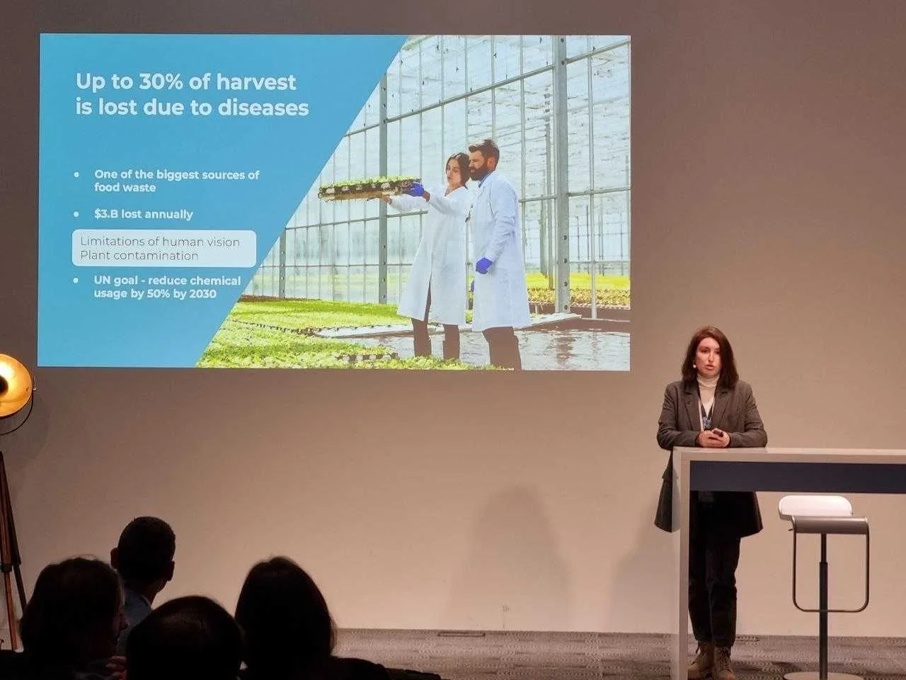 A woman giving a presentation in front of a projected slide about harvest loss and plant contamination in a greenhouse setting. The slide shows two people in lab coats inside a greenhouse with plants.