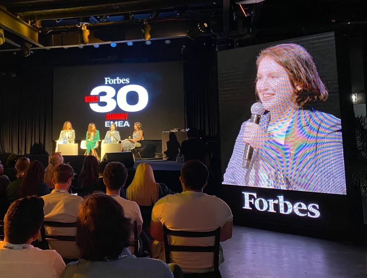 Panel discussion at the Forbes Under 30 Summit, with four speakers on stage, three women and one man, and an audience watching. A large screen shows a woman speaking into a microphone.