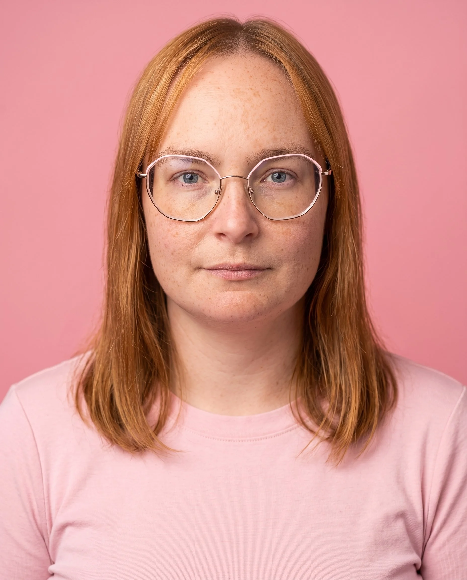 A woman with red hair and glasses, wearing a pink shirt, standing against a pink background.