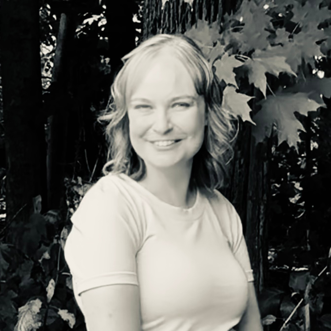 A woman with shoulder-length wavy hair smiling outdoors in front of large leafy plants.