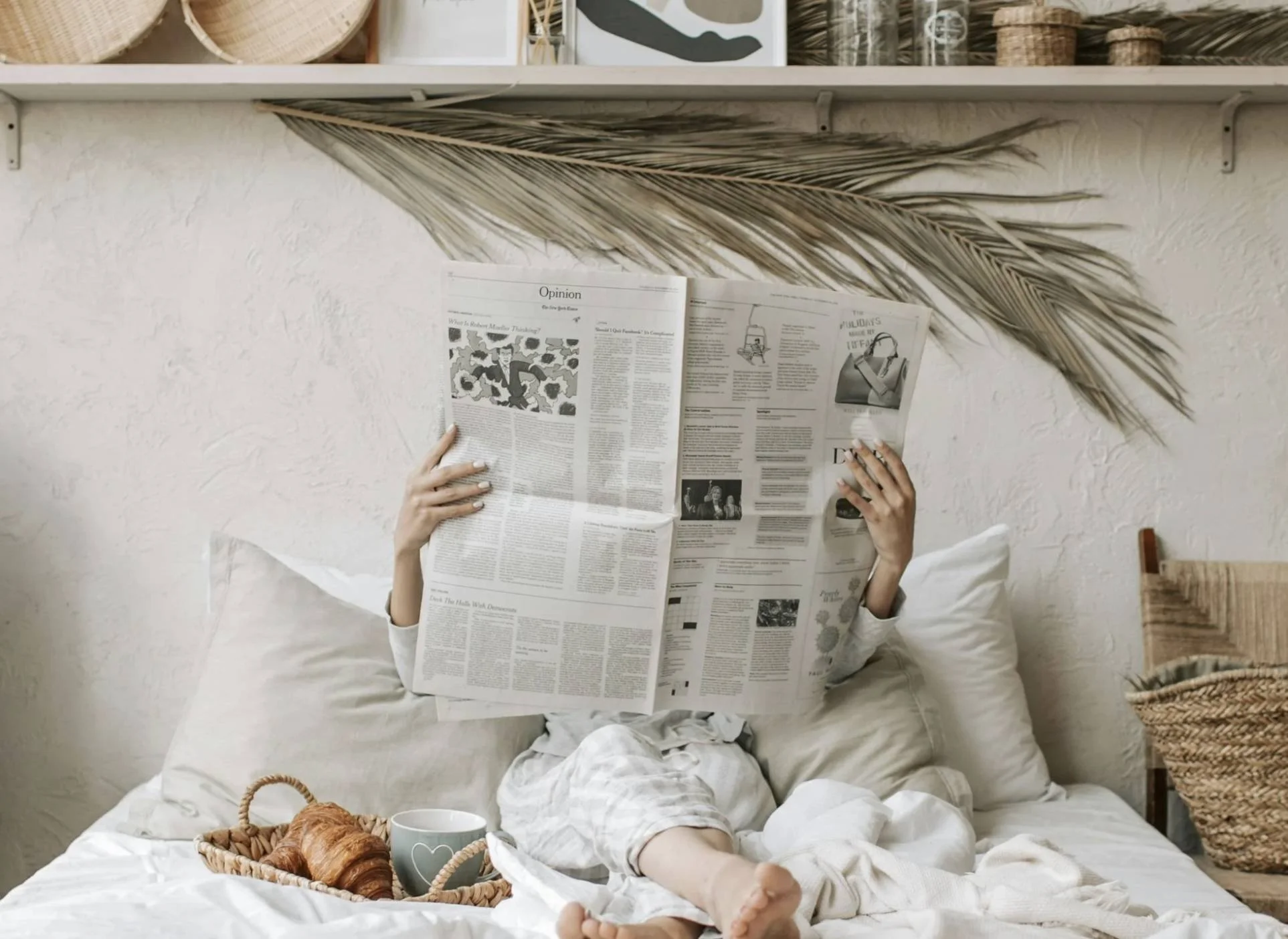Person lying on bed, reading a newspaper, with a basket of croissants and a mug nearby, in a cozy, well-decorated bedroom.