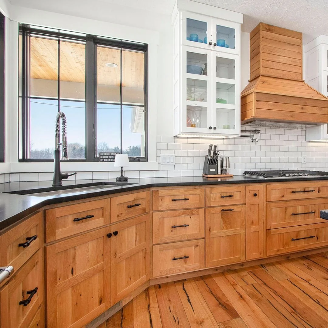 Kitchen with wooden cabinets, black countertop, white tile backsplash, window above the sink, and a built-in stove. There is a glass-front cabinet and a wooden range hood.