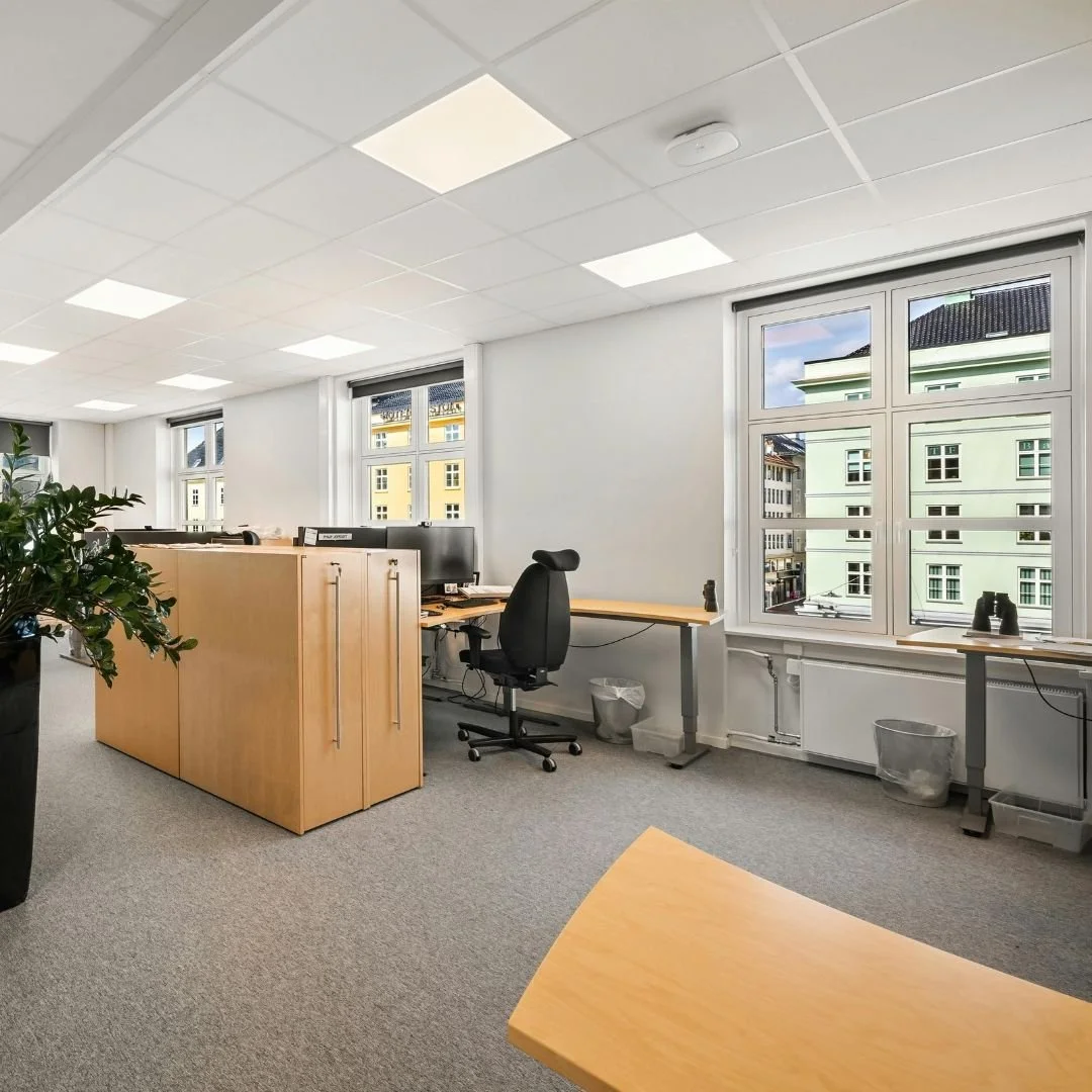 An empty office room with a large window, an ergonomic black office chair, a wooden desk with a computer, and a potted plant. Outside the window, neighboring buildings are visible.