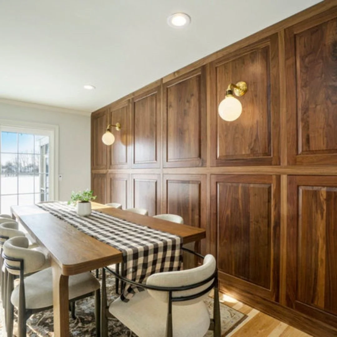 Dining room with wooden paneled wall, white chairs around a wooden table with a checkered table runner, potted plant centerpiece, and wall-mounted light fixtures, near a window with a view of the outside.