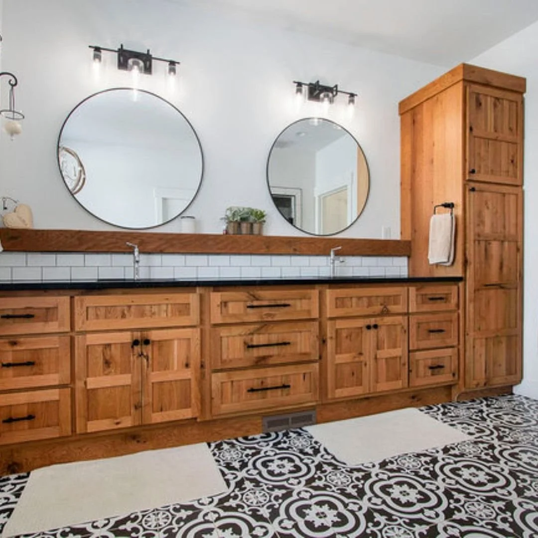 Bathroom vanity with two round mirrors, wooden cabinets, and black countertops, with patterned black and white tile floor.