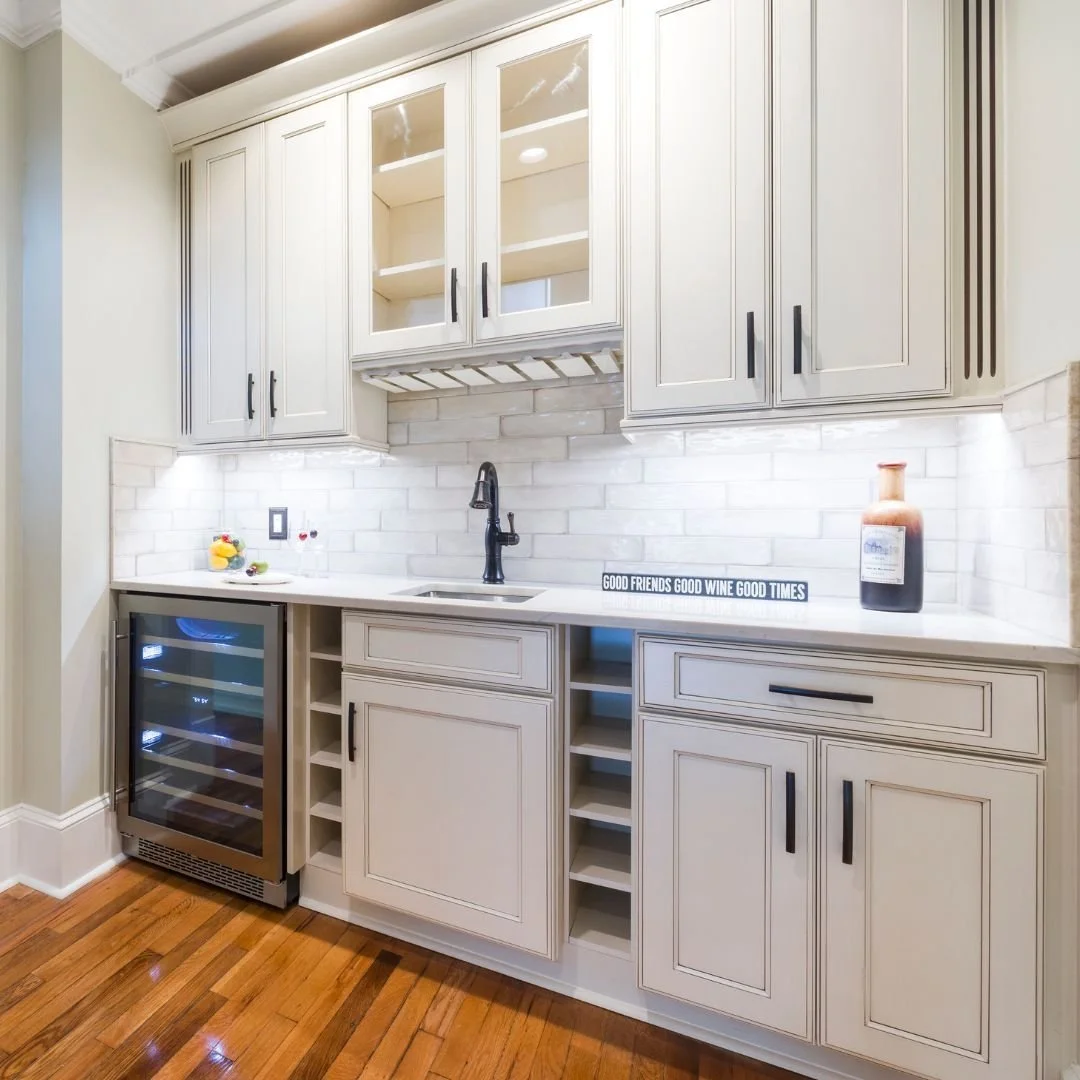 Modern kitchen with white cabinets, a wine cooler, open wine rack, a black faucet, and a white backsplash. Decor includes a small sign reading "Good friends good wine good times" and a wine bottle.