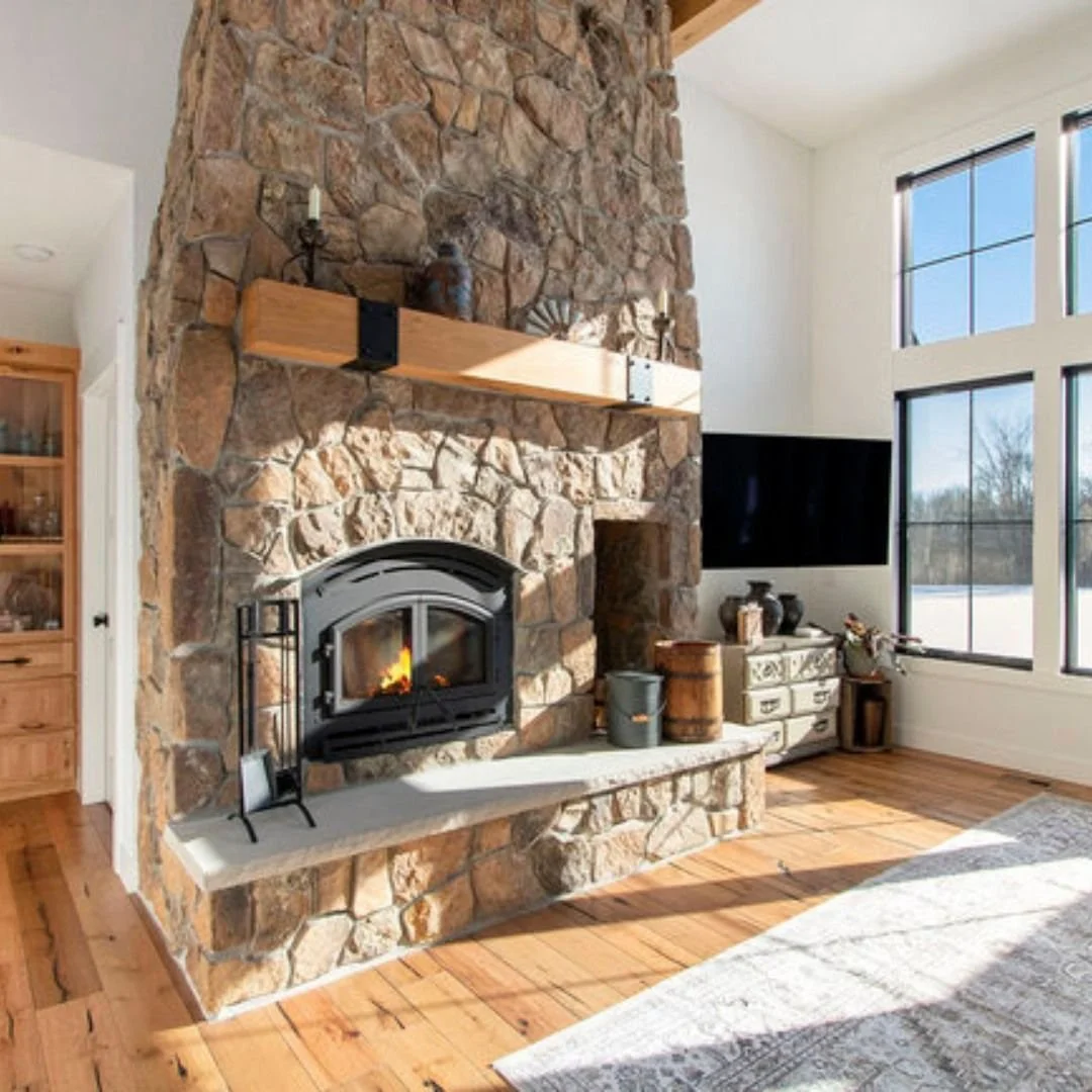 Living room with a large stone fireplace, a flat-screen TV, and hardwood flooring, with sunlight coming through big windows.