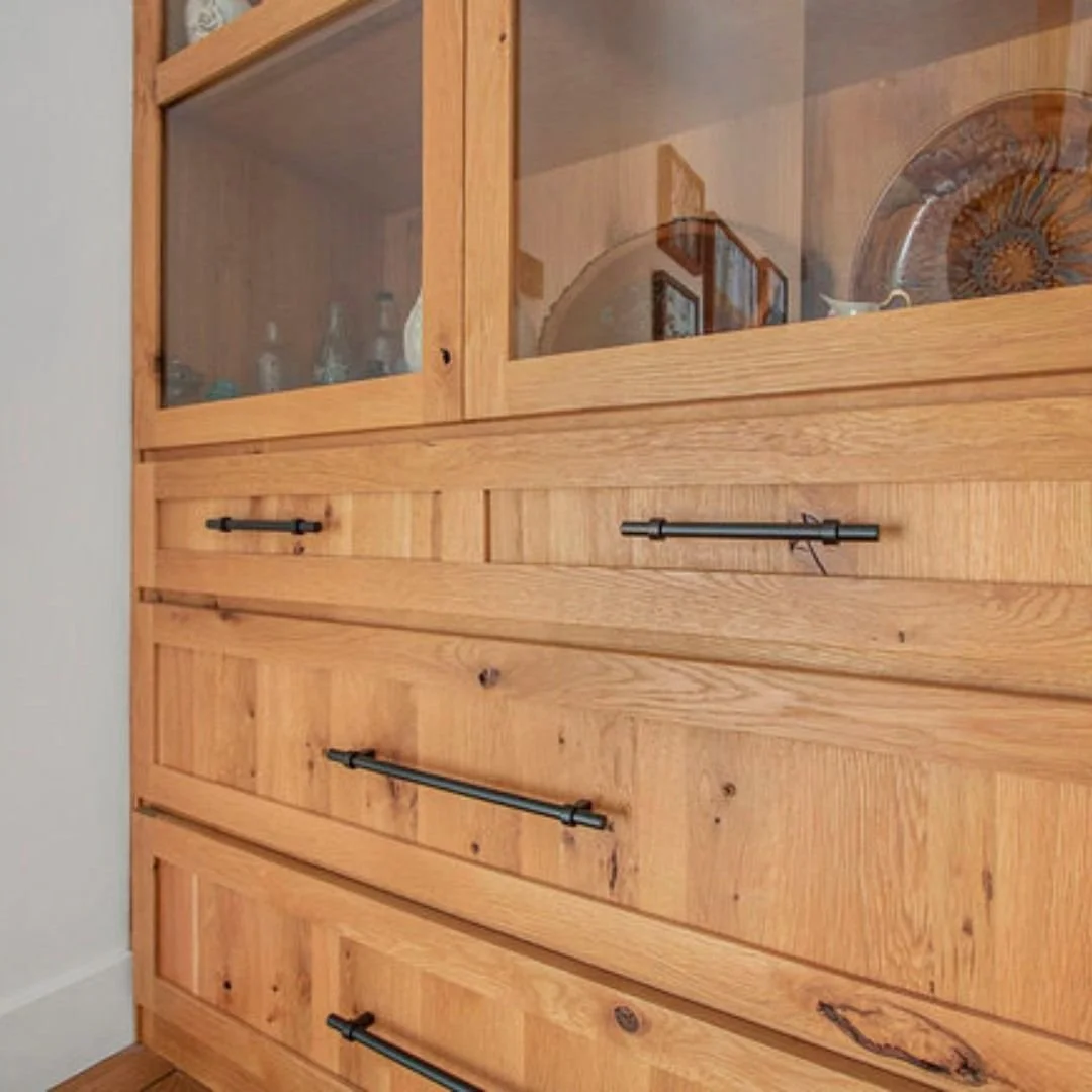 A wooden dining room hutch with glass doors on top and four drawers below, each with black metal handles. Inside the glass doors, decorative plates and other dishware can be seen.