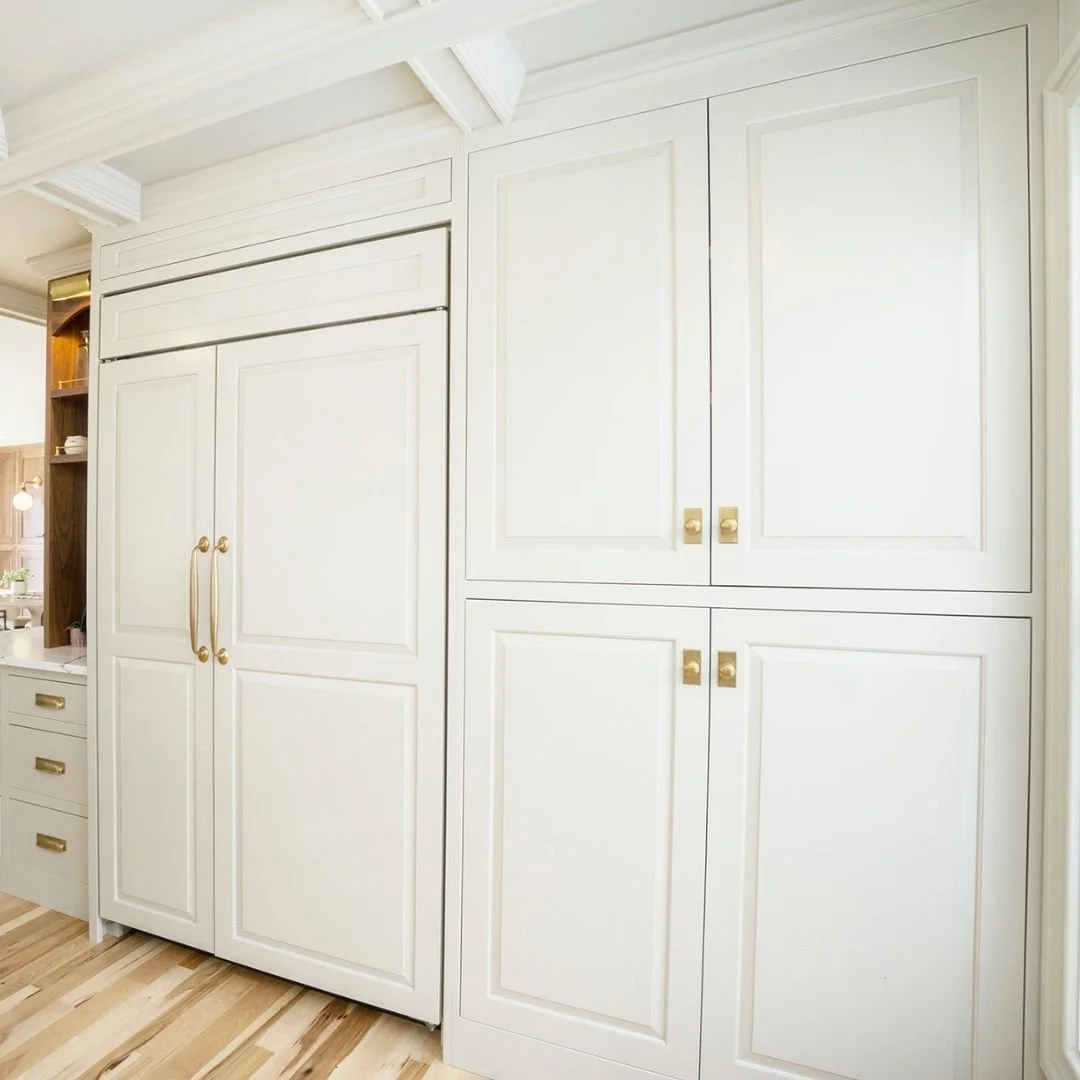 White built-in kitchen cabinets with gold handles and trim, adjacent to a small white dresser with gold hardware, wooden floor, and an open shelving area on the left.