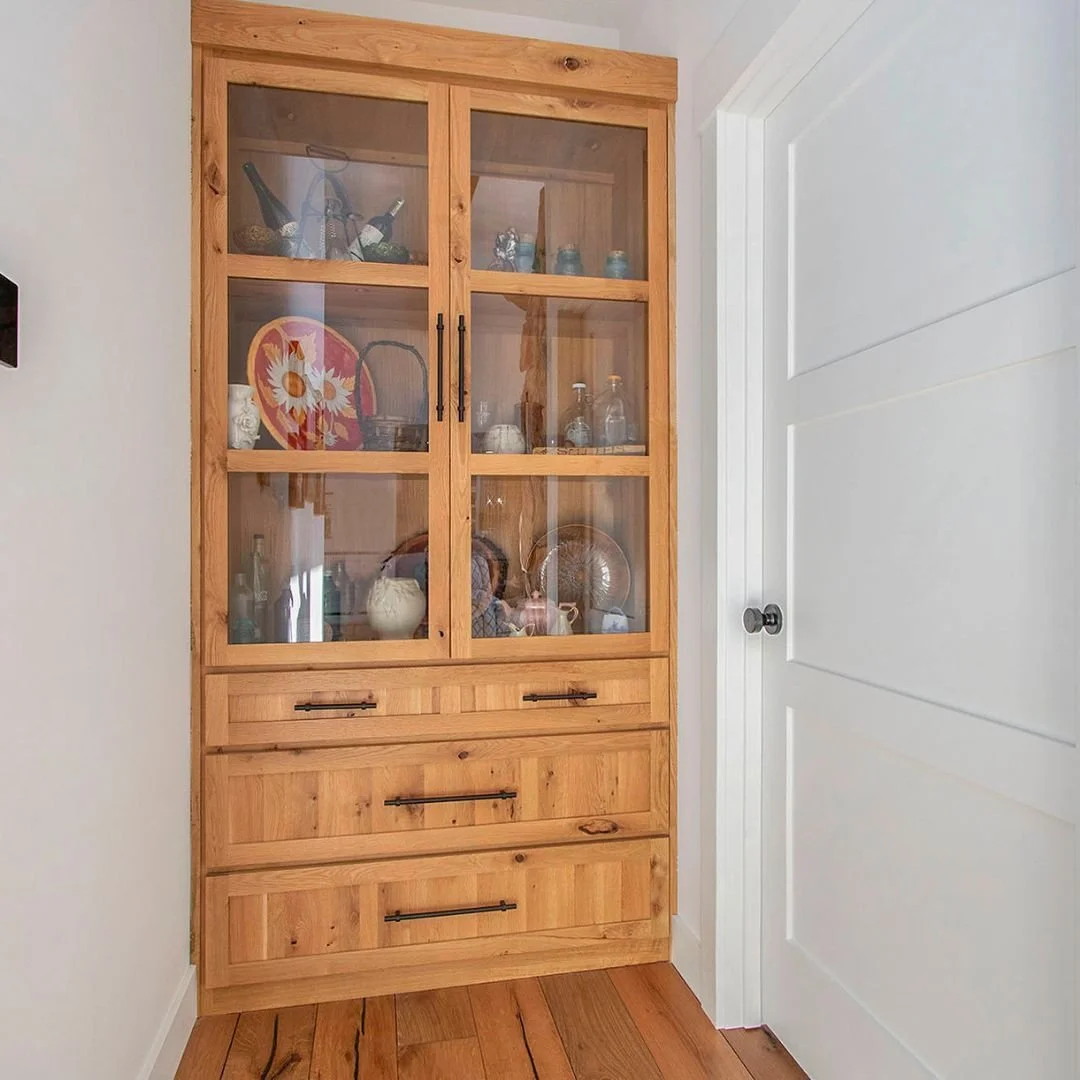 Wooden display cabinet with glass doors containing decorative items, bottles, and ceramics, placed in a corner next to a white wall and door.