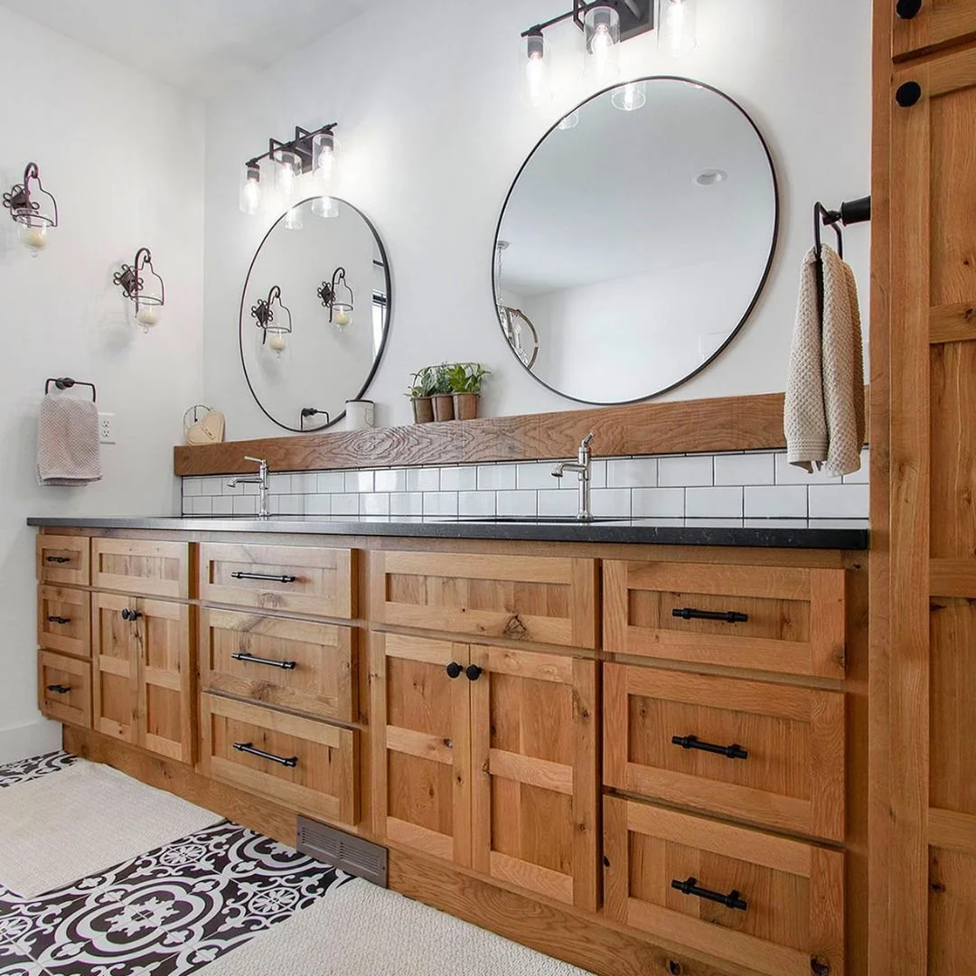 A bathroom vanity with a wooden cabinet, two round mirrors, black hardware, and a black countertop. Wall-mounted light fixtures above the mirrors, with candles. Small plants and decorative items on the counters. Towel racks with beige towels, white tiled backsplash, and patterned black-and-white floor tiles.