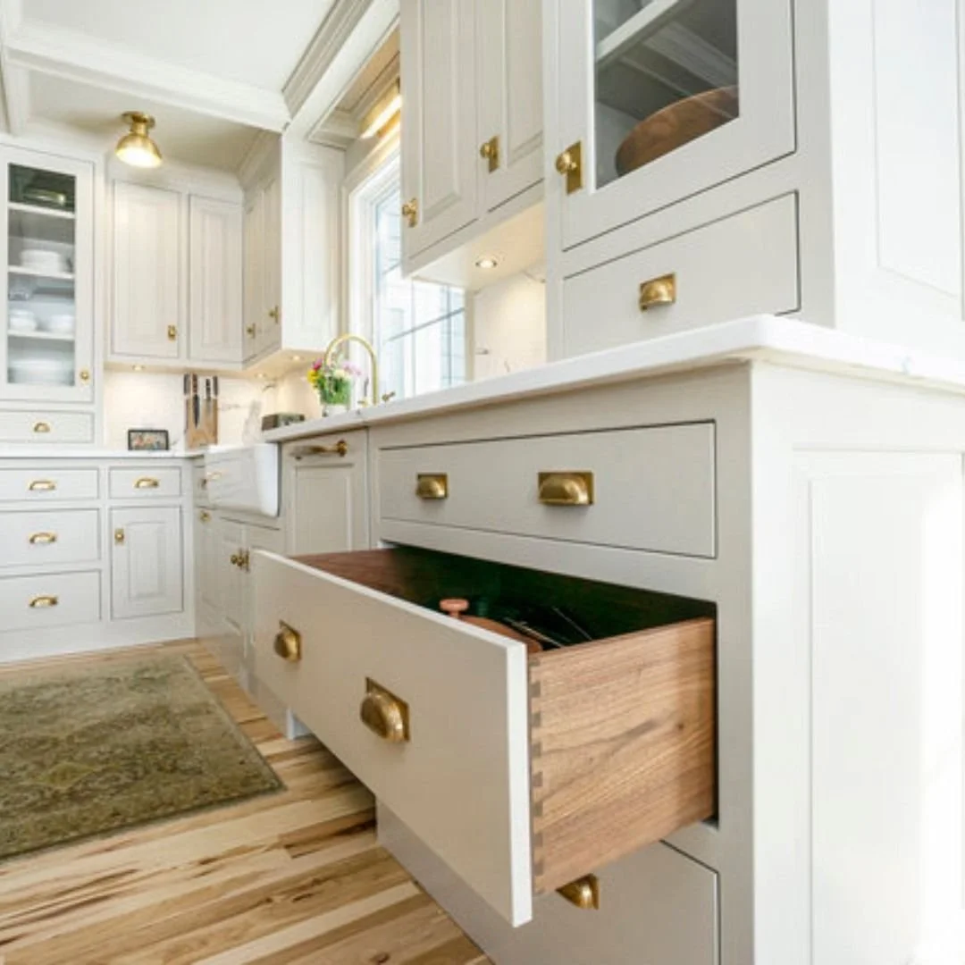 A bright, white kitchen with gold hardware, open shelves with dishes, and a partially open drawer showing wooden storage inside.