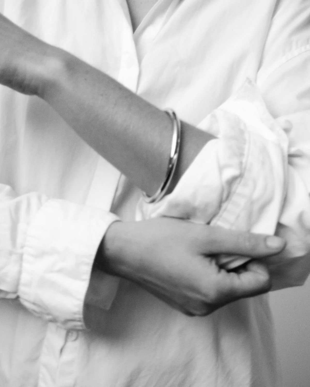 Close-up black and white photo showing a person wearing a white shirt and bracelets on their arm, with hands crossed over their body.