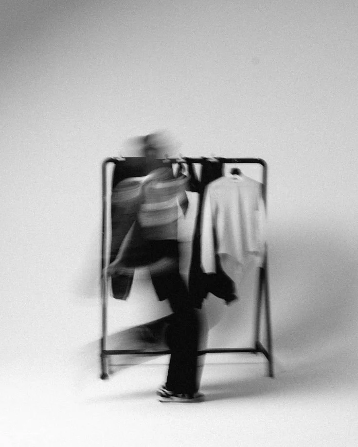 Blurred person shopping for clothes at a clothing rack with hanging garments in a plain, monochrome setting.
